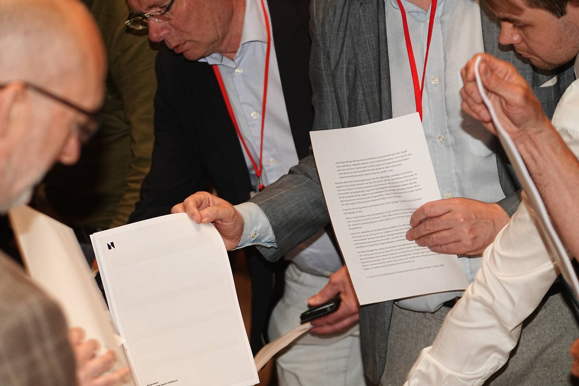 Flere personer holder og ser på dokumenter. En mann i grå dress holder et dokument med tekst og en logo øverst. En annen mann, iført en mørk jakke, ser på et annet dokument. Rundt personene er det andre mennesker som også er engasjert i dokumentene. Det er røde snorer festet rundt halsen på noen av personene. (Bildebeskrivelsen er laget av en KI-tjeneste)