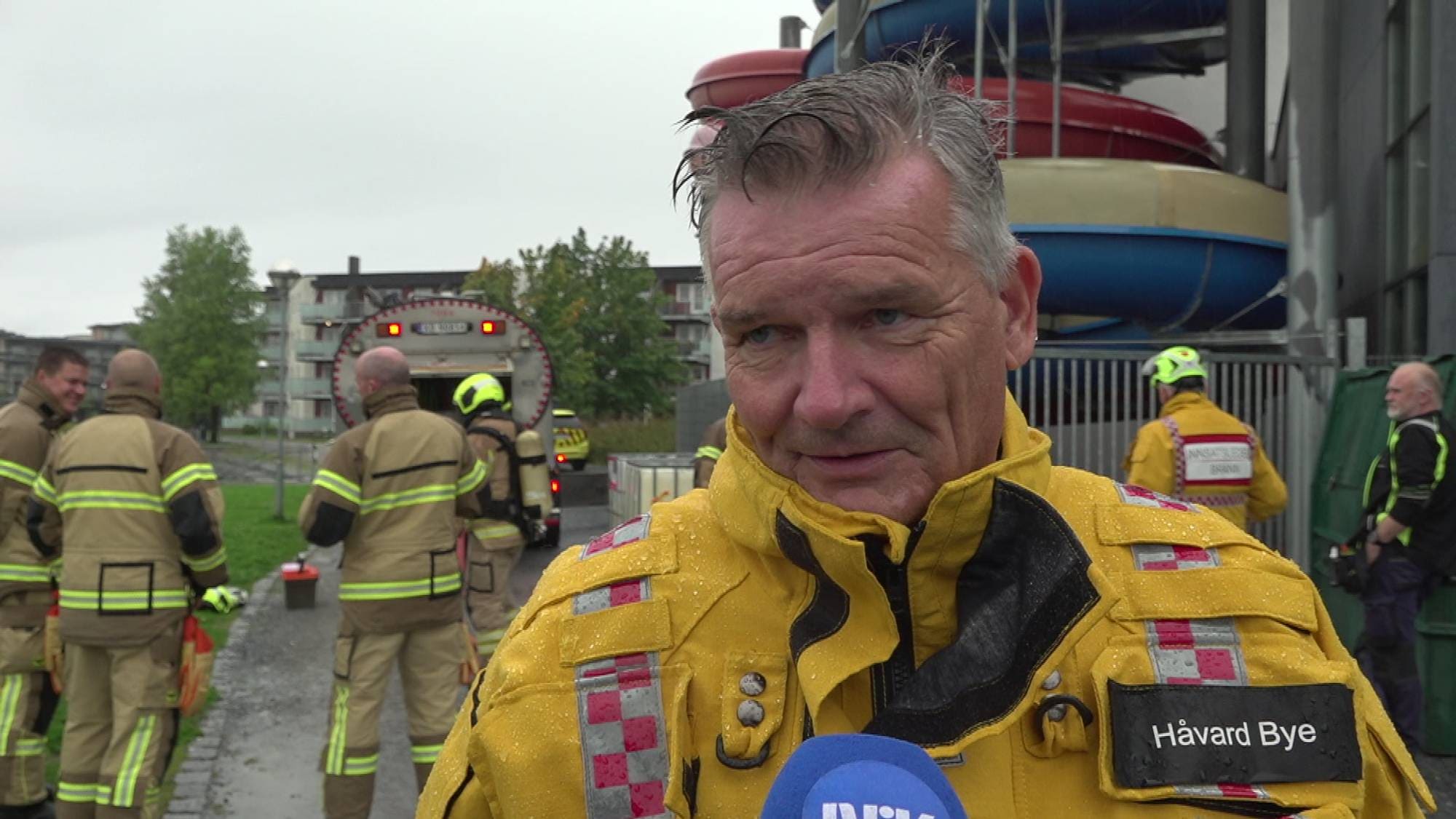 En mann i gul uniform, med brannvesenet bak seg.