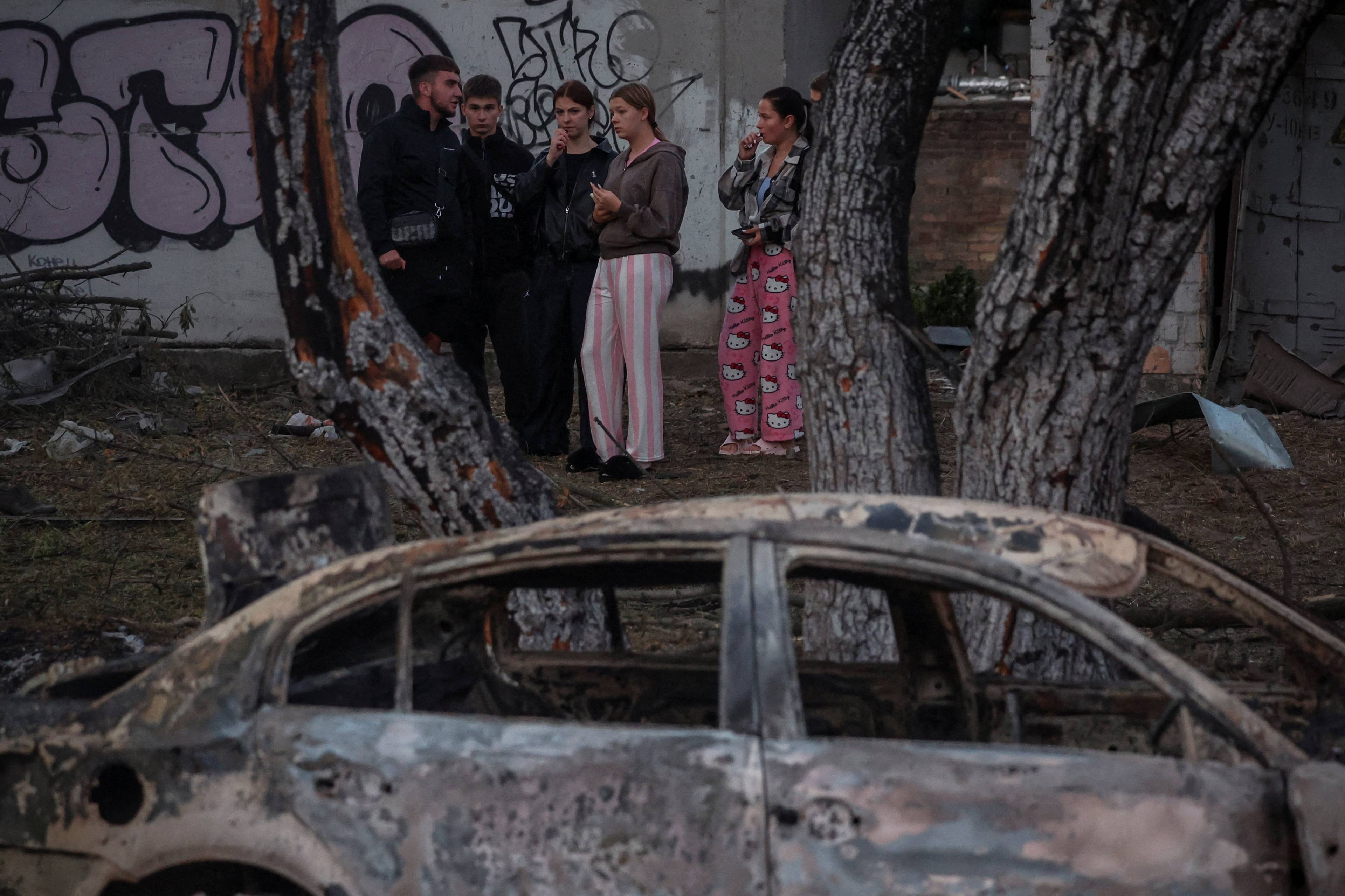 Fem ungdommer står ved en graffitivegg bak et utbrent bilvrak. (Bildebeskrivelsen er laget av en KI-tjeneste)