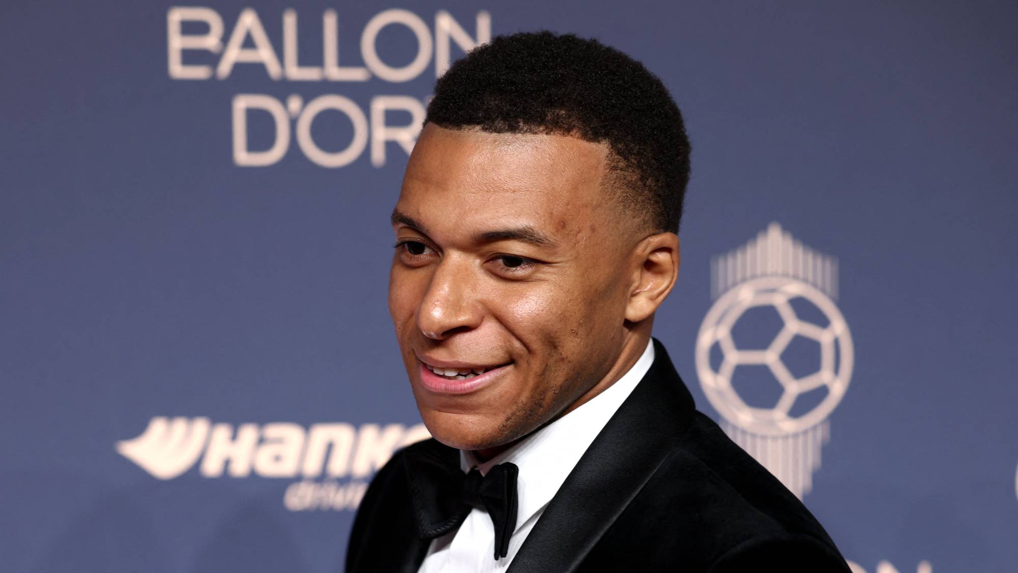 Fotballspilleren Kylian Mbappé i dress og sløyfe foran en Ballon d'Or-bakgrunn. (Bildebeskrivelsen er laget av en KI-tjeneste)