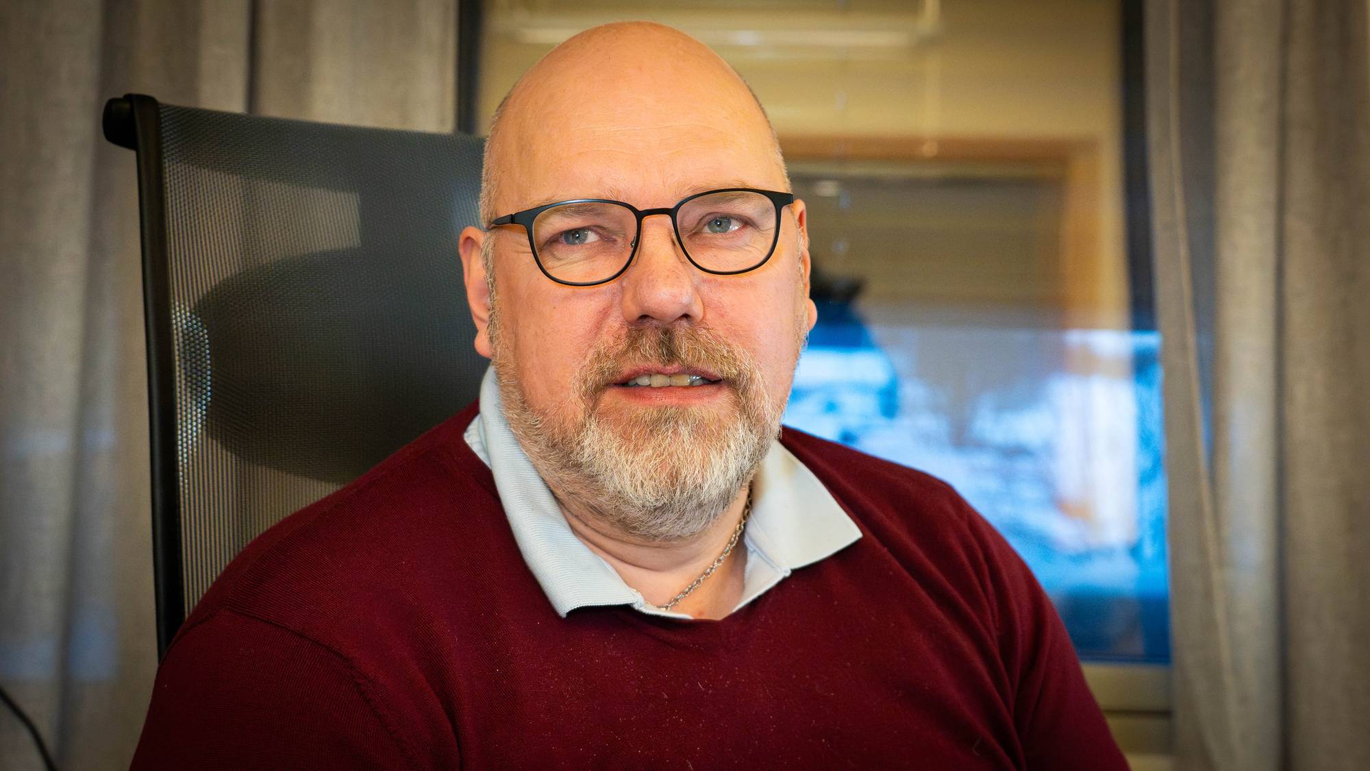 En mann sitter foran et vindu i et rom med gardiner. Han har på seg en burgunder genser og er delvis skjult bak en stol. Omgivelsene ser ut til å være kontoraktige, med noen uskarpe elementer i bakgrunnen. Mannen har en alvorlig mine og ser mot kameraet. (Bildebeskrivelsen er laget av en KI-tjeneste)