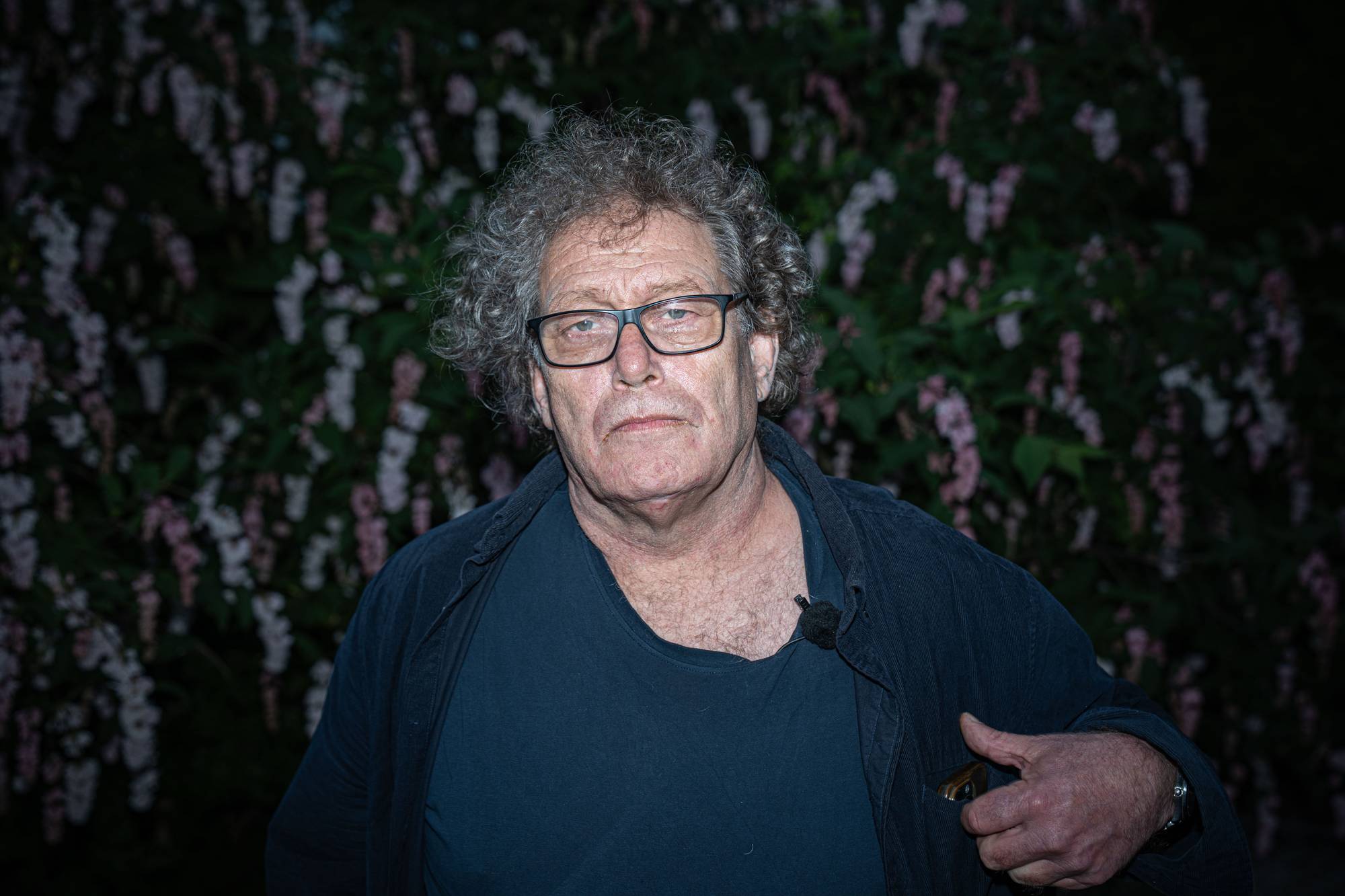 En mann med grått, krøllete hår og briller står foran en blomstrende busk. Han har på seg en mørkegrå jakke over en mørkeblå t-skjorte. Mannen ser mot kameraet. En mikrofon er festet til t-skjorten hans. (Bildebeskrivelsen er laget av en KI-tjeneste)