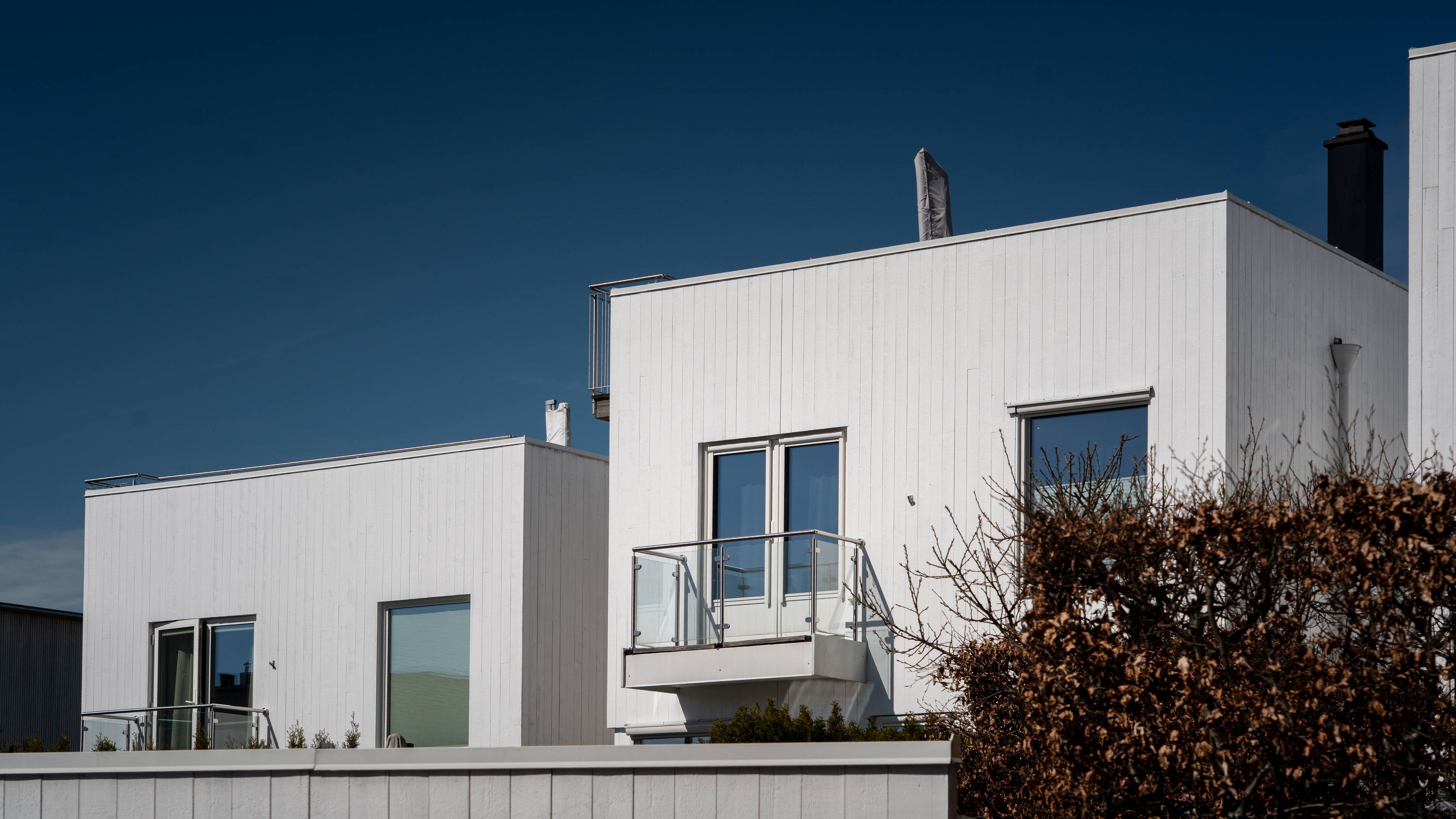 To hvite trehus med moderne arkitektur står under en mørkeblå himmel. En av bygningene har en balkong med glassrekkverk og doble dører som fører ut til den. Foran husene er et lavt, hvitt trepanelgjerde. En tørket busk med brune blader dekker en del av bildet. (Bildebeskrivelsen er laget av en KI-tjeneste)
