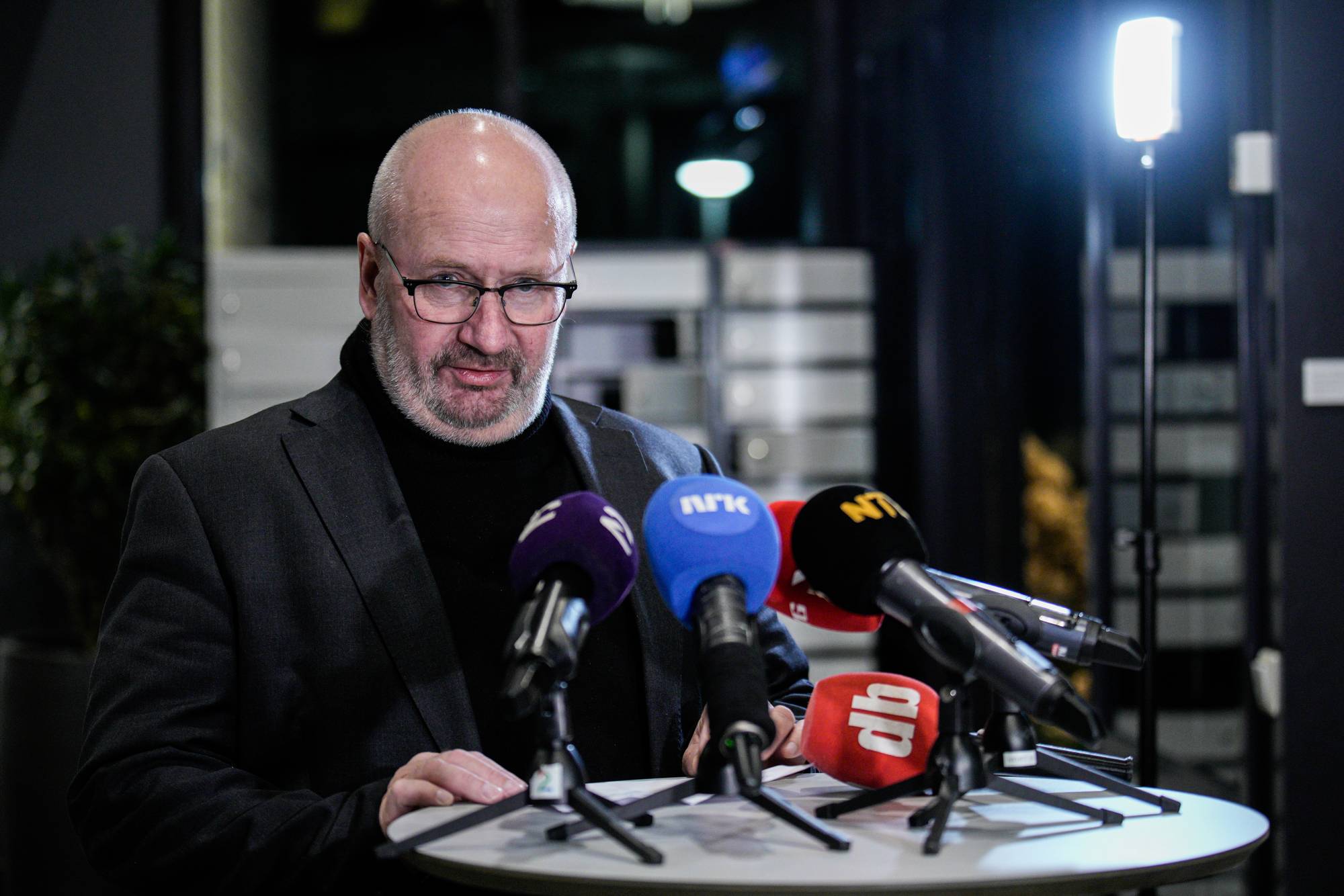 En mann med briller og dress sitter bak et bord. Flere mikrofoner med logoer fra NRK og Dagbladet er plassert foran ham. Bak mannen er det svakt opplyste omgivelser med en lampe i bakgrunnen.