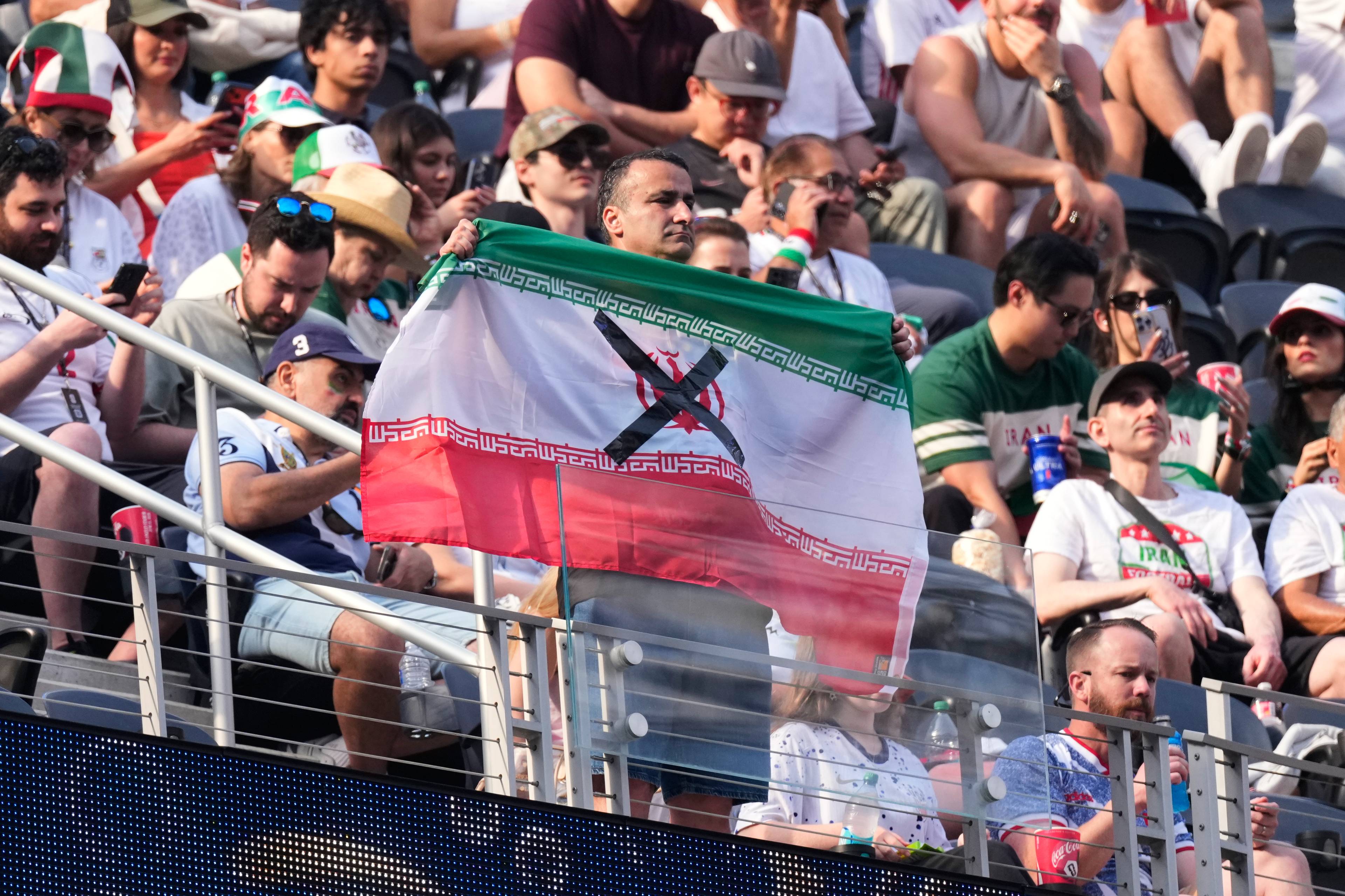 En mann holder opp et iransk flagg på tribunen. Mannen er omgitt av tilskuere på en stadion. Flagget har et sort kryss med en rød form under på den hvite stripen. (Bildebeskrivelsen er laget av en KI-tjeneste)