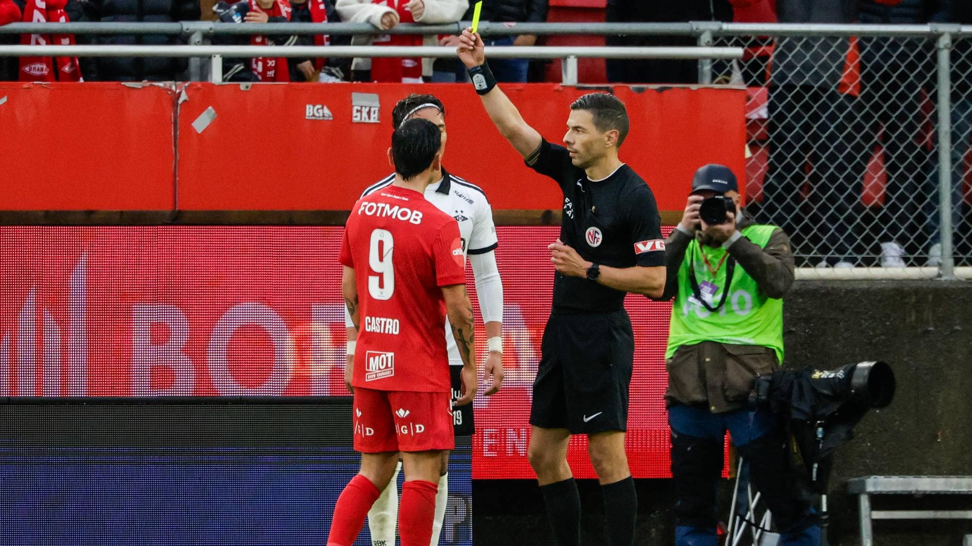 Dommer gir gult kort til en spiller på fotballbanen. Fotograf står ved siden av banen. (Bildebeskrivelsen er laget av en KI-tjeneste)