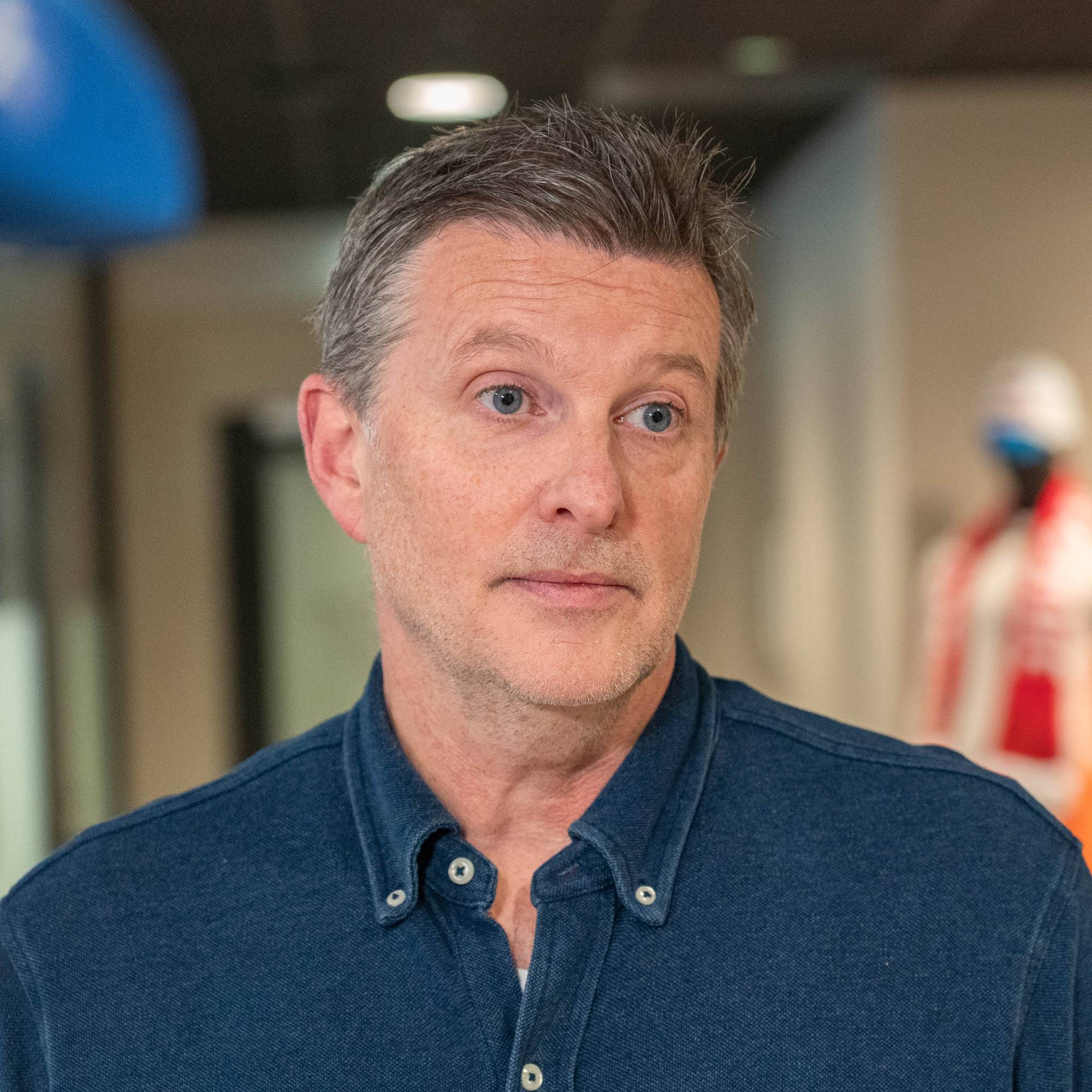 En mann står i et kontormiljø iført en blå skjorte. I bakgrunnen kan man se en mannequindukke kledd i sportsutstyr. Rommet har dempet belysning og noen planter er synlige. Det er en profesjonell setting preget av arbeid og samarbeid.