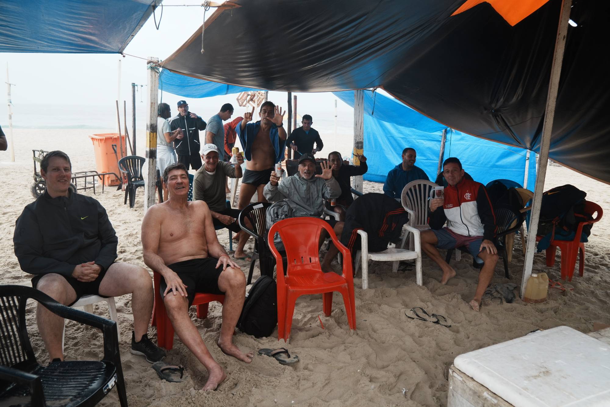 En gruppe menn sitter under et presenning-telt på en strand og slapper av med drikke. (Bildebeskrivelsen er laget av en KI-tjeneste)