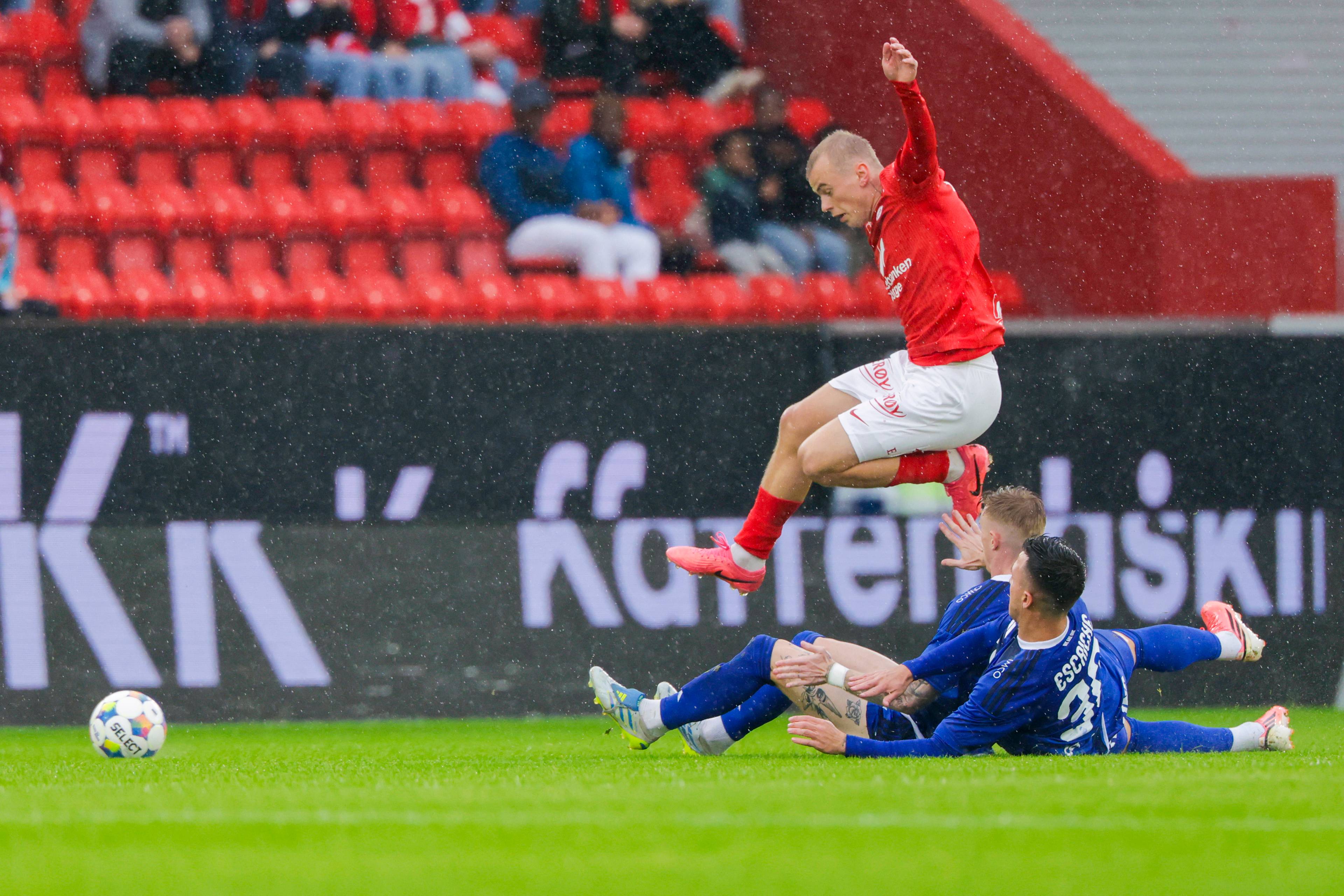 Fotballspiller i rødt hopper over en motspiller i blått i en duell på gressbanen. (Bildebeskrivelsen er laget av en KI-tjeneste)