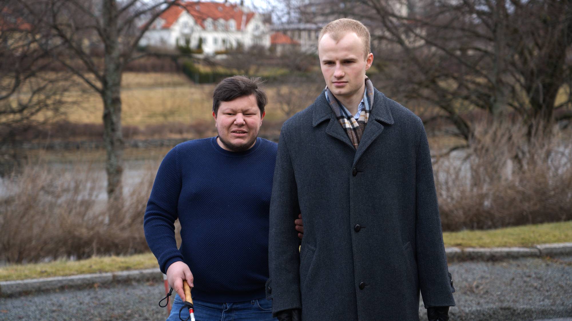 En blind mann går sammen med sin støttekontakt, foran en elv. Den blinde holder staven sin, mens han holder i armen til støttekontakten.
