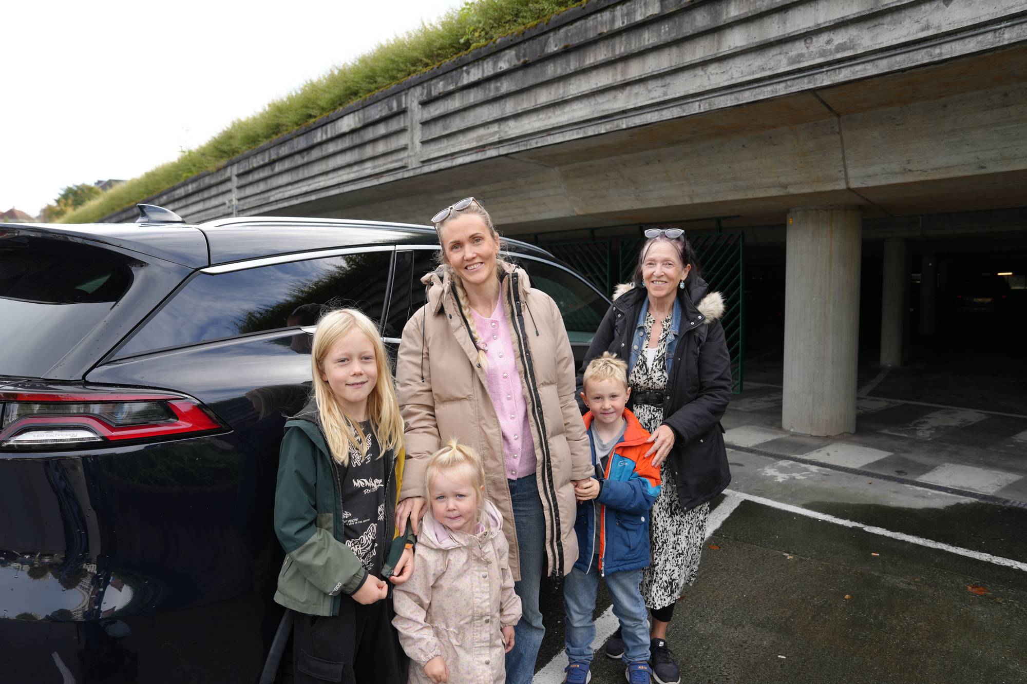 To kvinner og tre barn står ved en parkert bil utenfor en parkeringsgarasje. (Bildebeskrivelsen er laget av en KI-tjeneste)