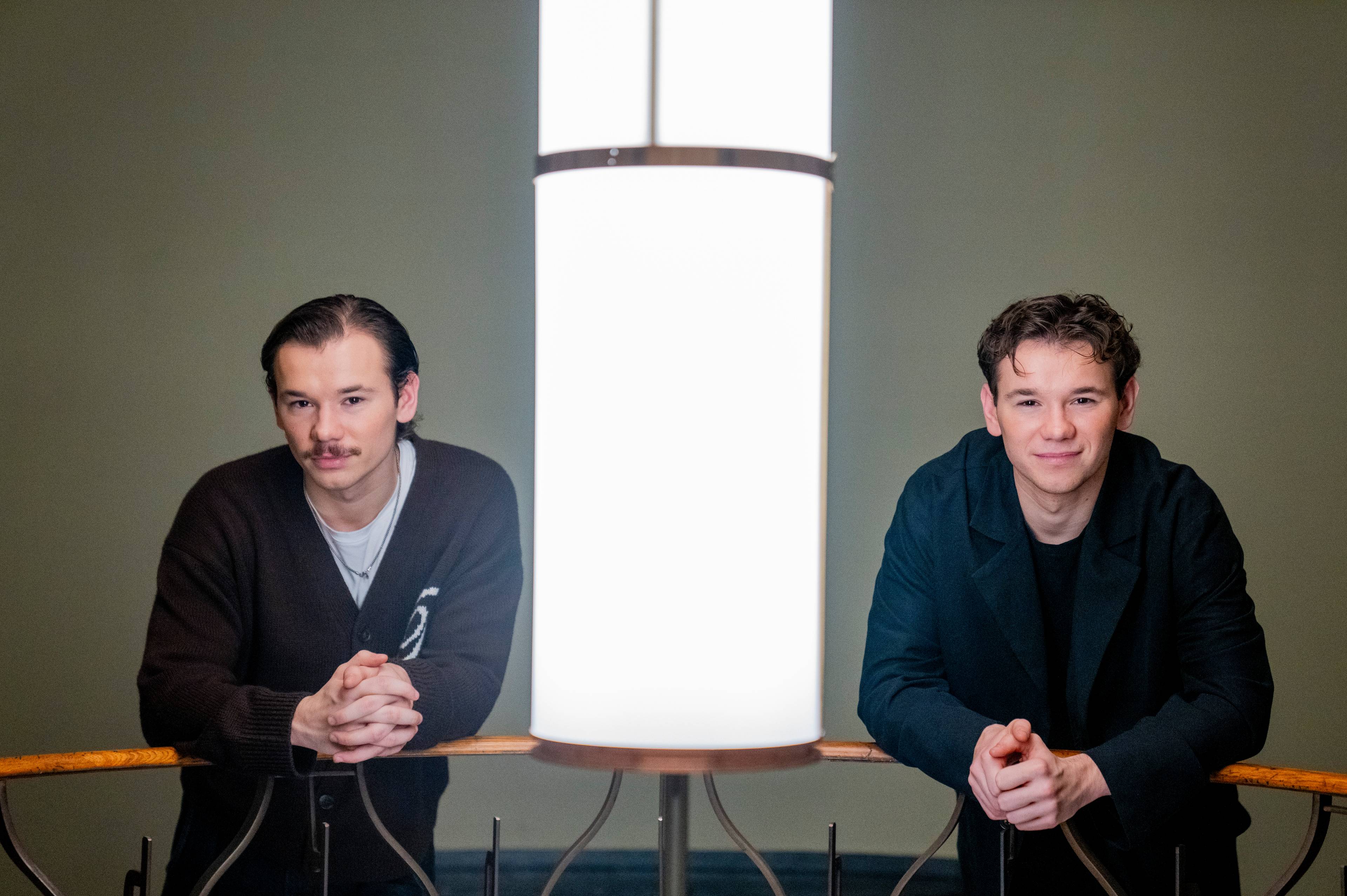 To unge menn lener seg mot et rekkverk. Begge ser mot kamera og smiler. Mellom dem står en høy, sylindrisk lampe. (Bildebeskrivelsen er laget av en KI-tjeneste)