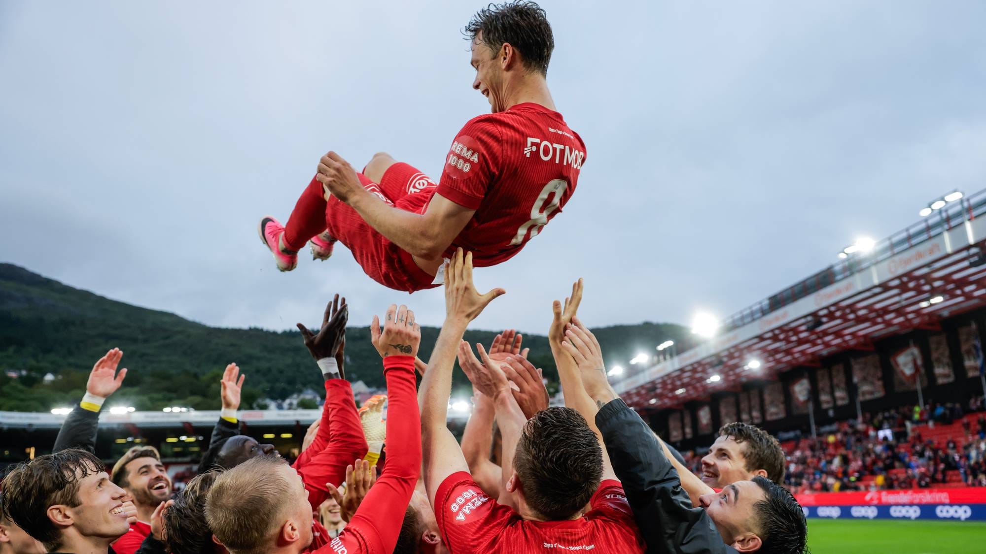 Fotballspillere i røde drakter løfter en lagkamerat opp i luften på en stadion. 