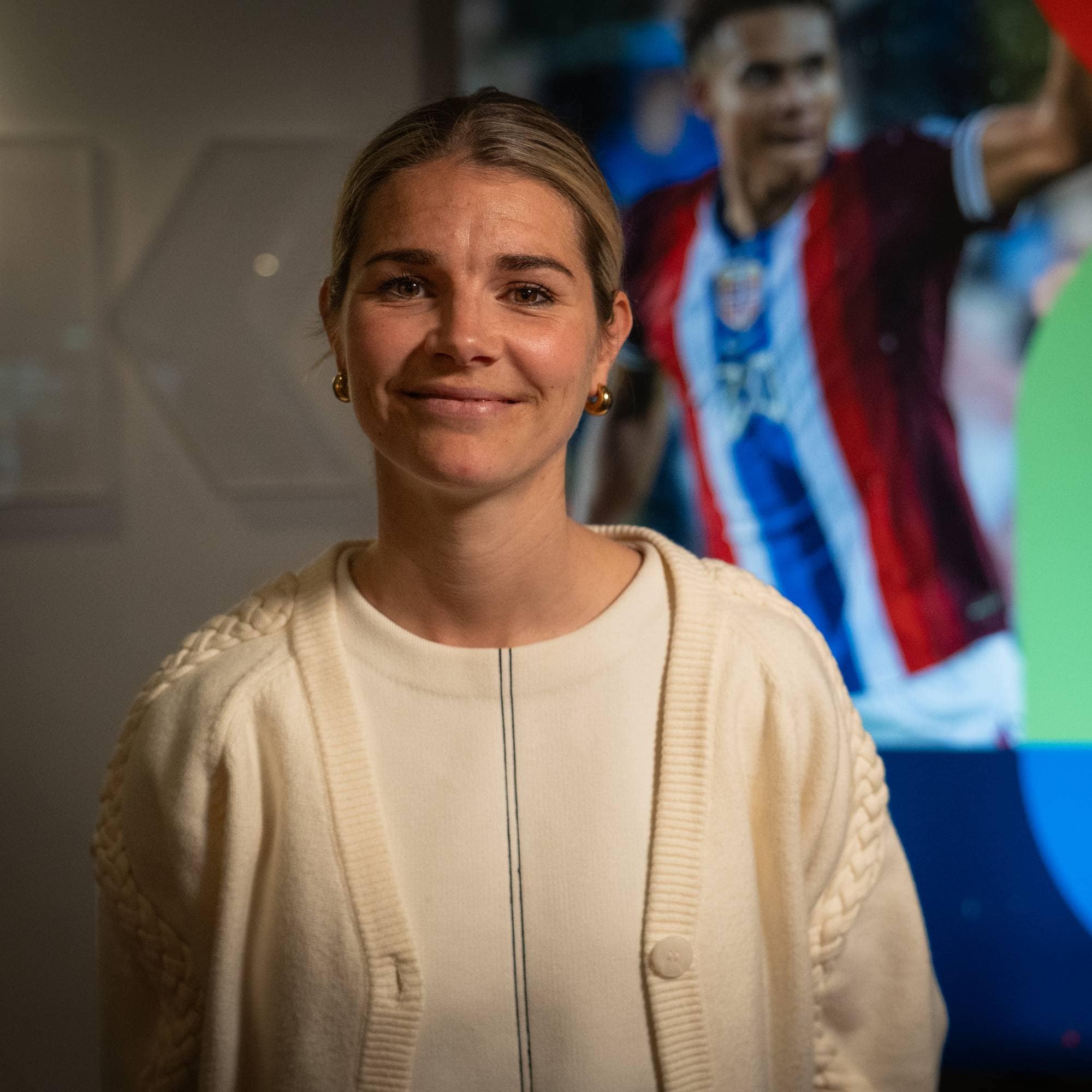 En kvinne med blondt hår smiler mot kamera. Bak henne er et bilde av en fotballspiller i en drakt. Kvinnen har på seg en hvit genser og en beige cardigan. Bakgrunnen er mørk og abstrakt, med fargede sirkler. (Bildebeskrivelsen er laget av en KI-tjeneste)