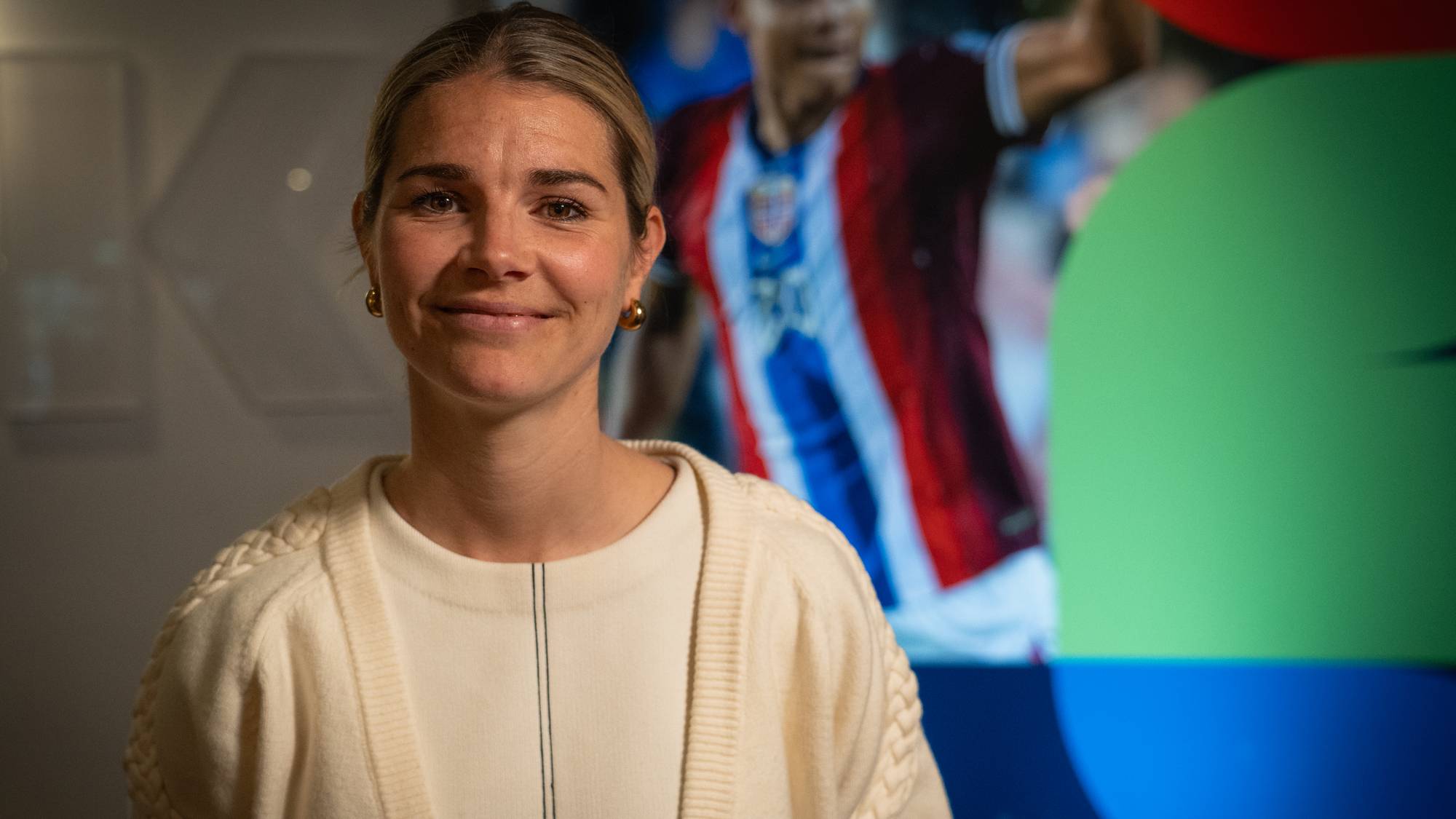 En kvinne med blondt hår smiler mot kamera. Bak henne er et bilde av en fotballspiller i en drakt. Kvinnen har på seg en hvit genser og en beige cardigan. Bakgrunnen er mørk og abstrakt, med fargede sirkler. (Bildebeskrivelsen er laget av en KI-tjeneste)