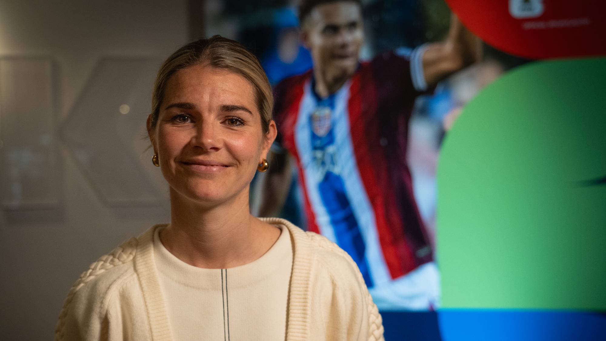 En kvinne med blondt hår smiler mot kamera. Bak henne er et bilde av en fotballspiller i en drakt. Kvinnen har på seg en hvit genser og en beige cardigan. Bakgrunnen er mørk og abstrakt, med fargede sirkler. (Bildebeskrivelsen er laget av en KI-tjeneste)