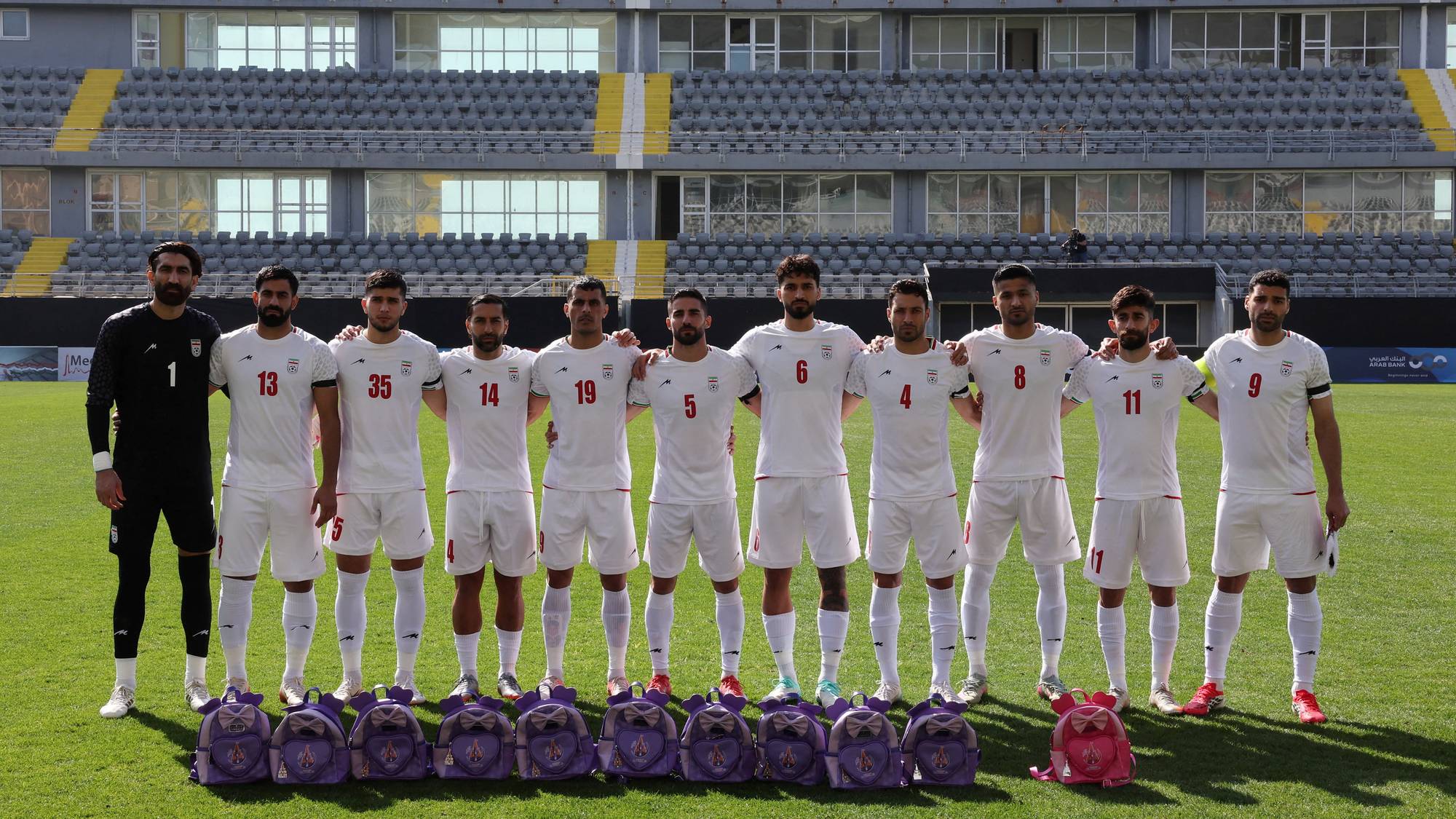Elve fotballspillere står på en gressbane. Alle har på seg hvite drakter med iranske symboler. Bakerst i bildet er en tribune med grå seter. Foran spillerne står tolv lilla barnebagger. (Bildebeskrivelsen er laget av en KI-tjeneste)