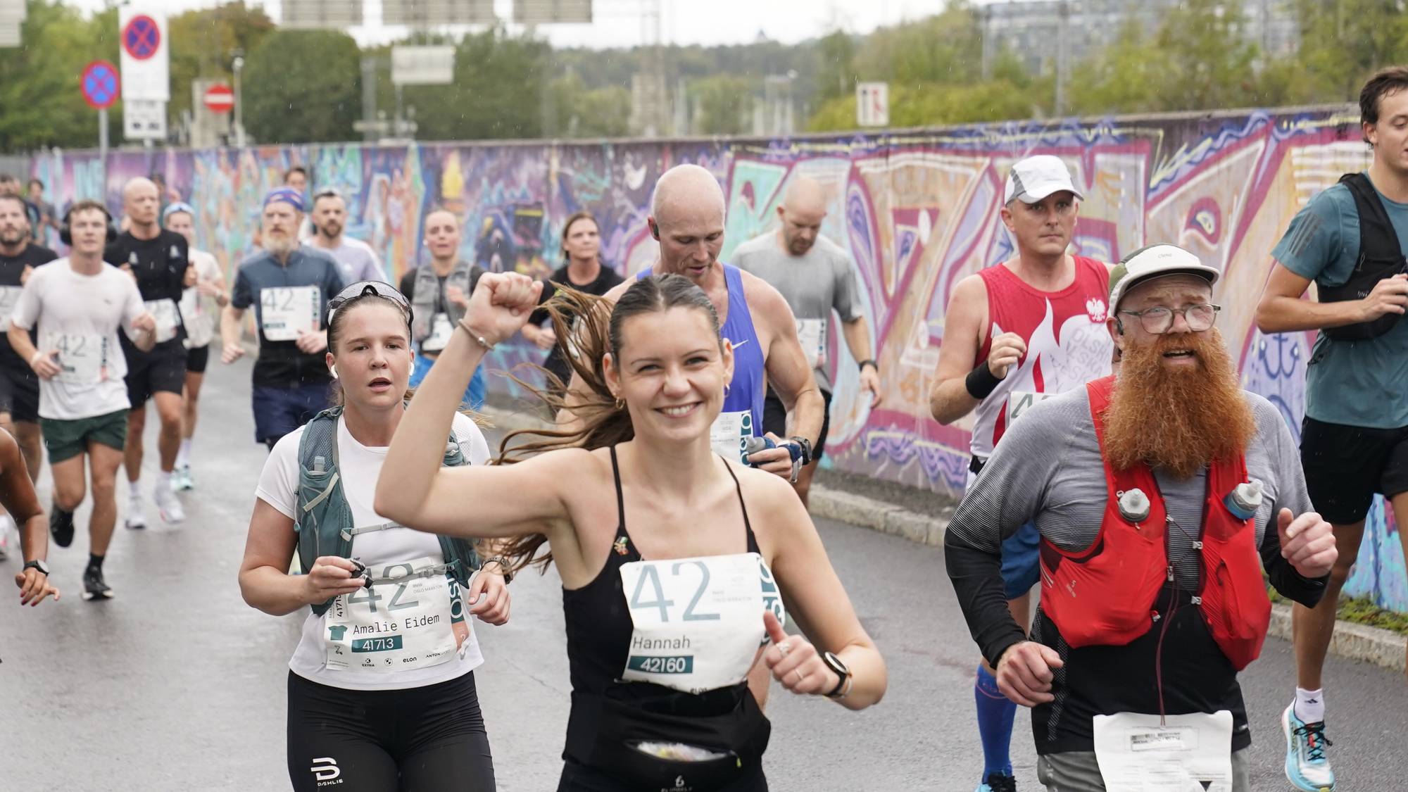 Deltakere løper Oslo maraton forbi en graffiti-vegg. En kvinne i forgrunnen løfter neven i jubel.