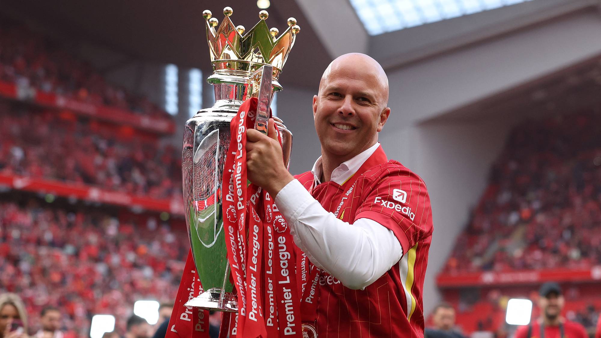 En mann i rød Liverpool-drakt holder Premier League-trofeet. Mannen smiler og ser mot kamera. I bakgrunnen er det publikum på en stadion. (Bildebeskrivelsen er laget av en KI-tjeneste)