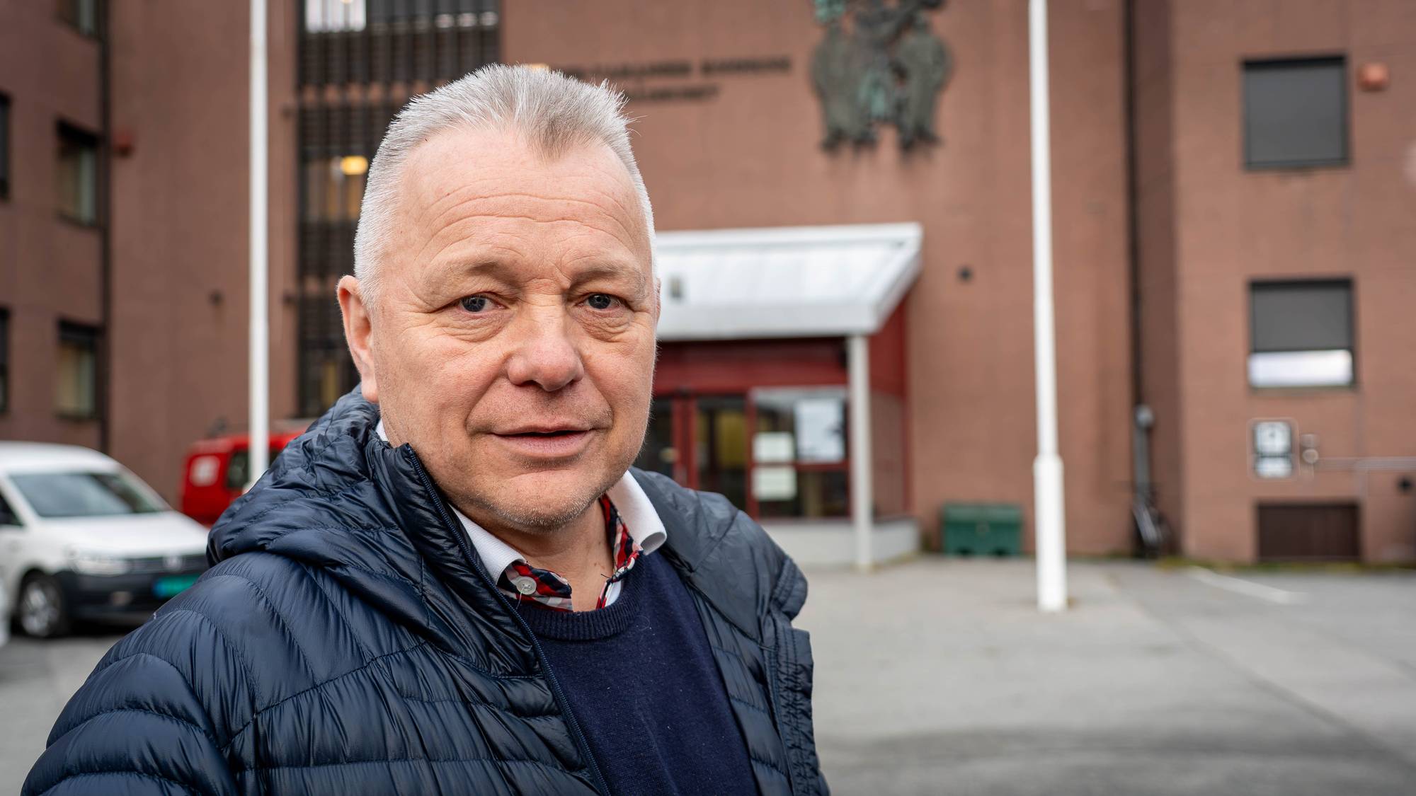 Mann står foran en bygning med et våpenskjold på veggen. Bakgrunnen viser inngangen til et bygg. (Bildebeskrivelsen er laget av en KI-tjeneste)