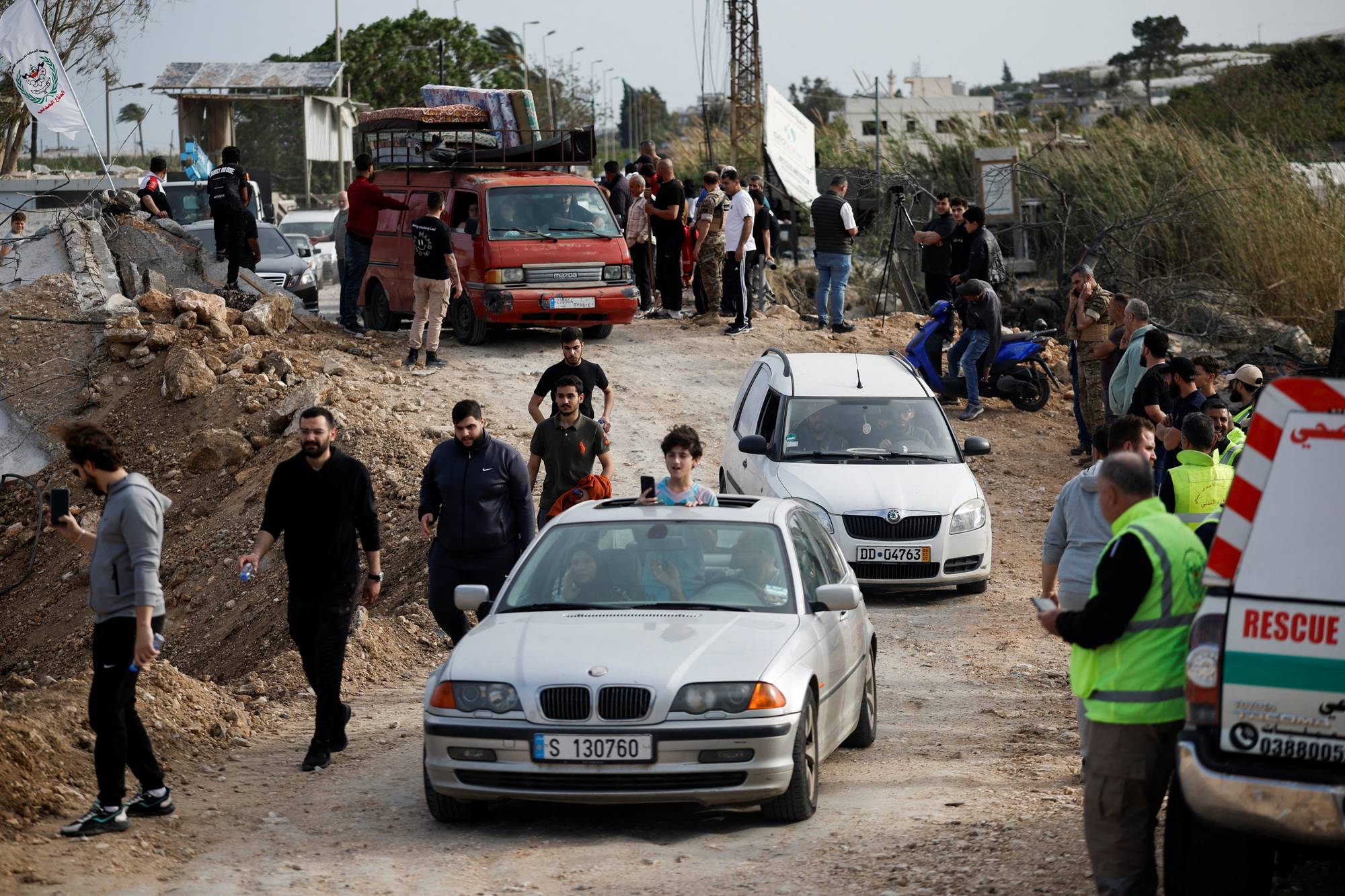 En gruppe mennesker står langs en grusvei der flere biler kjører. Noen er kledd i gule vester. En rød varebil med bagasje på taket er synlig. Det er ruiner og vegetasjon i bakgrunnen. (Bildebeskrivelsen er laget av en KI-tjeneste)