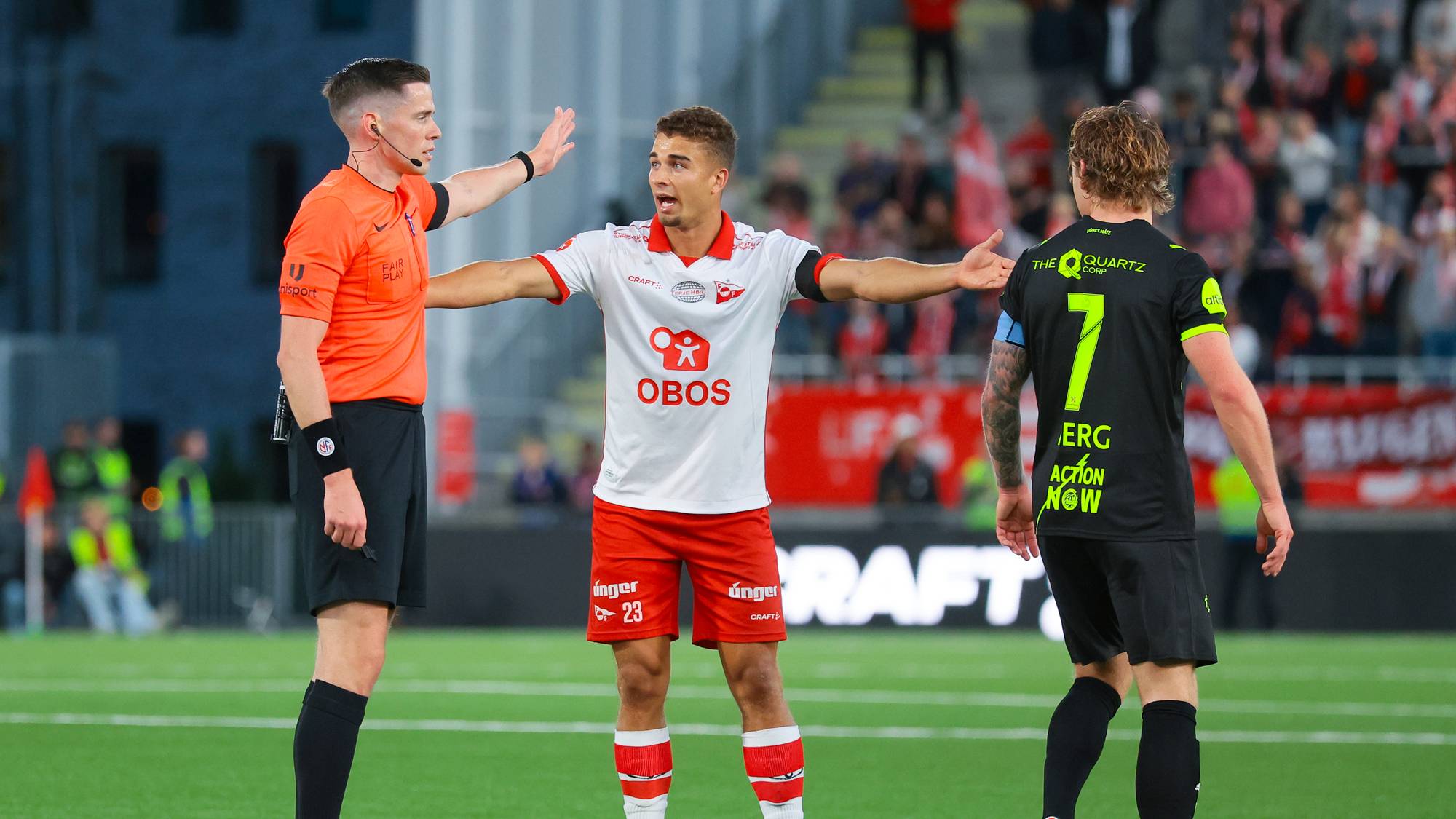En fotballdommer snakker med en spiller mens en annen spiller går forbi på en fotballbane. (Bildebeskrivelsen er laget av en KI-tjeneste)