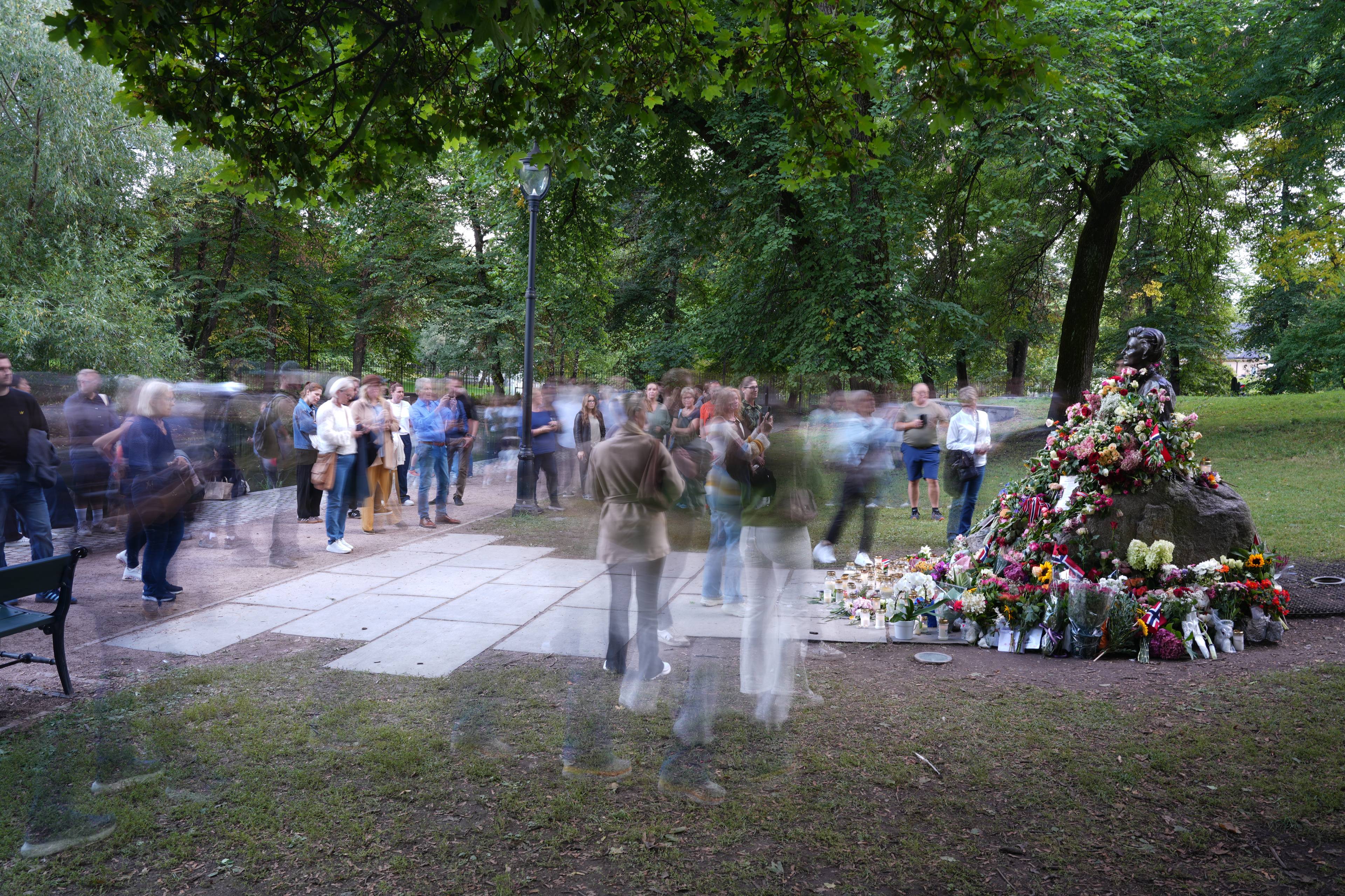 Folk samles ved en blomsterdekket statue i en park, delvis preget av lang lukkertid. (Bildebeskrivelsen er laget av en KI-tjeneste)