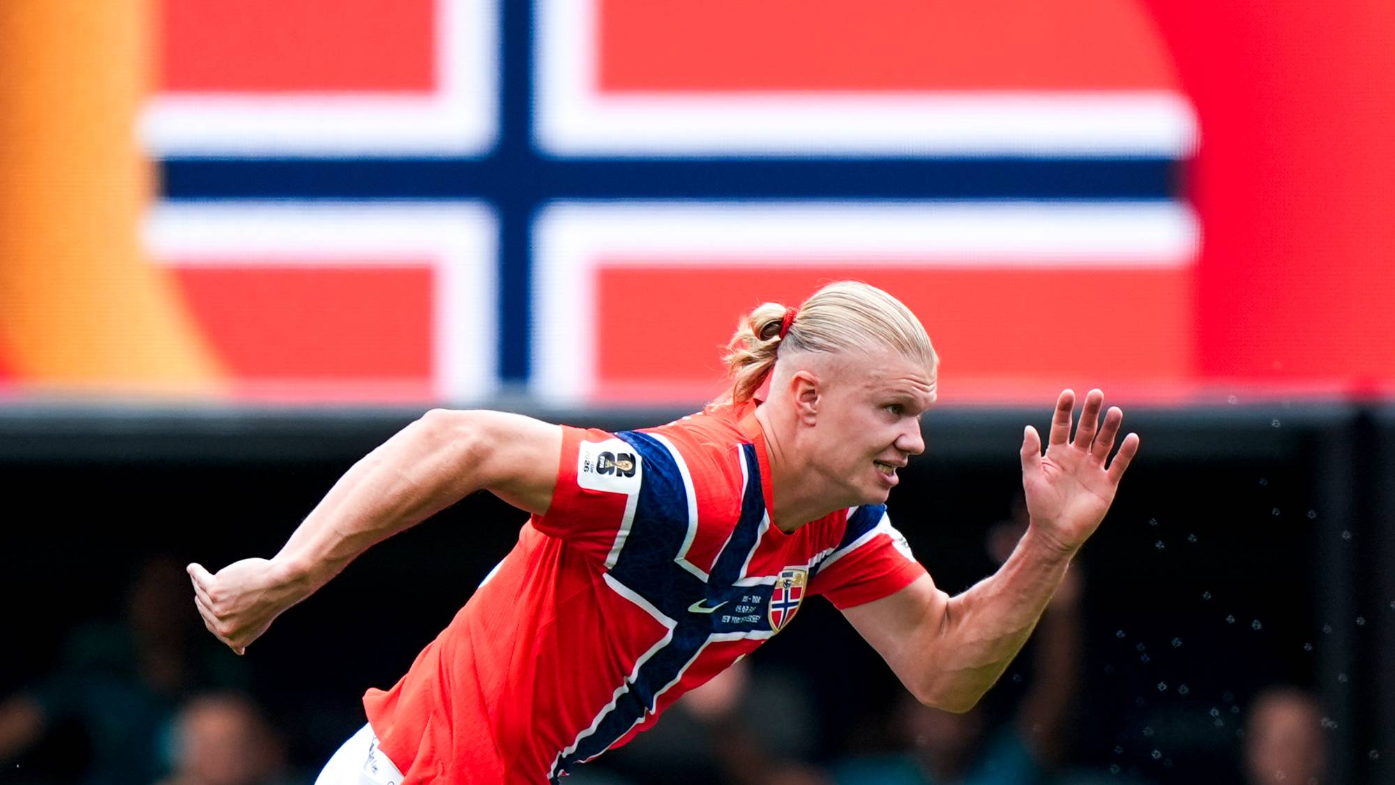 Erling Braut Haaland løper på en fotballbane foran et norsk flagg. (Bildebeskrivelsen er laget av en KI-tjeneste)