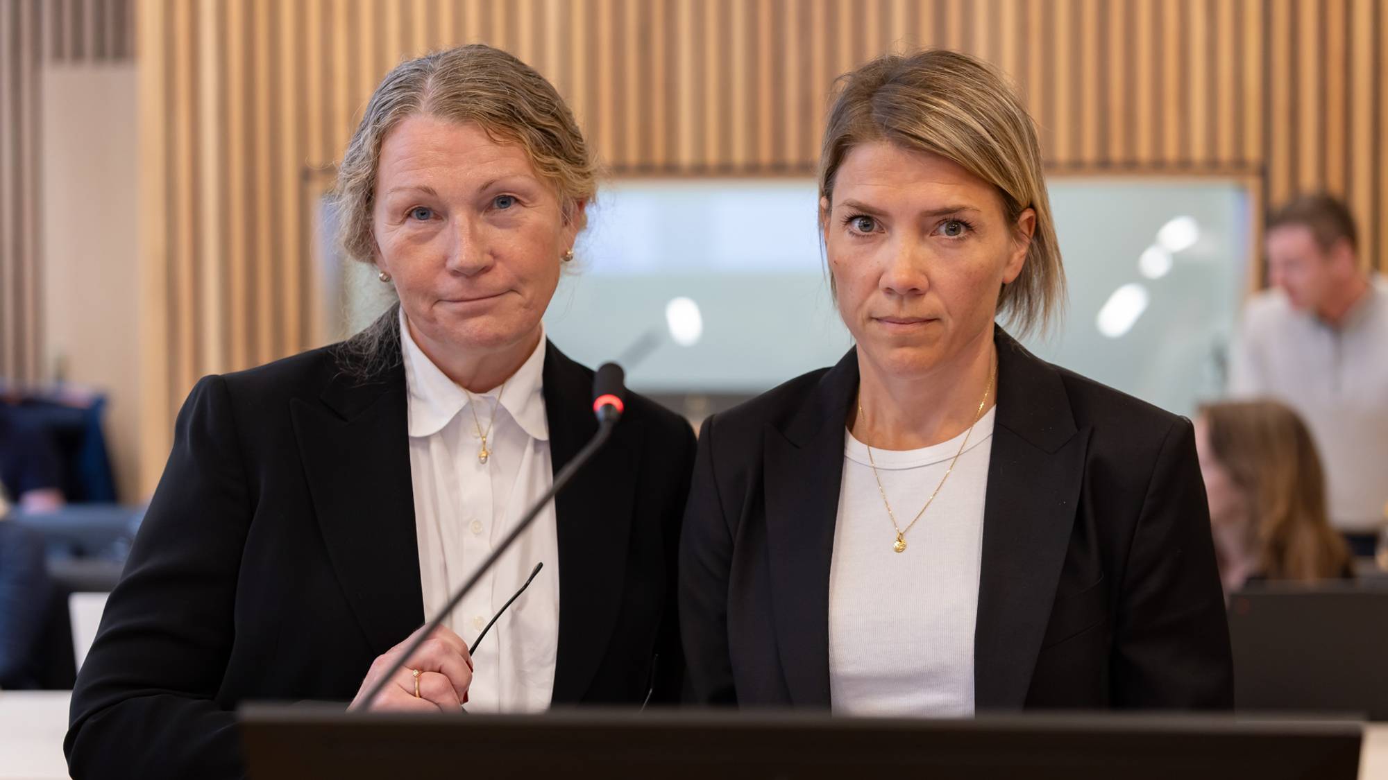To kvinner står ved en benk i en rettssal. De er begge iført svarte klær. De står ved en mikrofon og er klar til å gi sin vurdering av tiltalte for retten. I bakgrunnen kan vi se noen journalister
