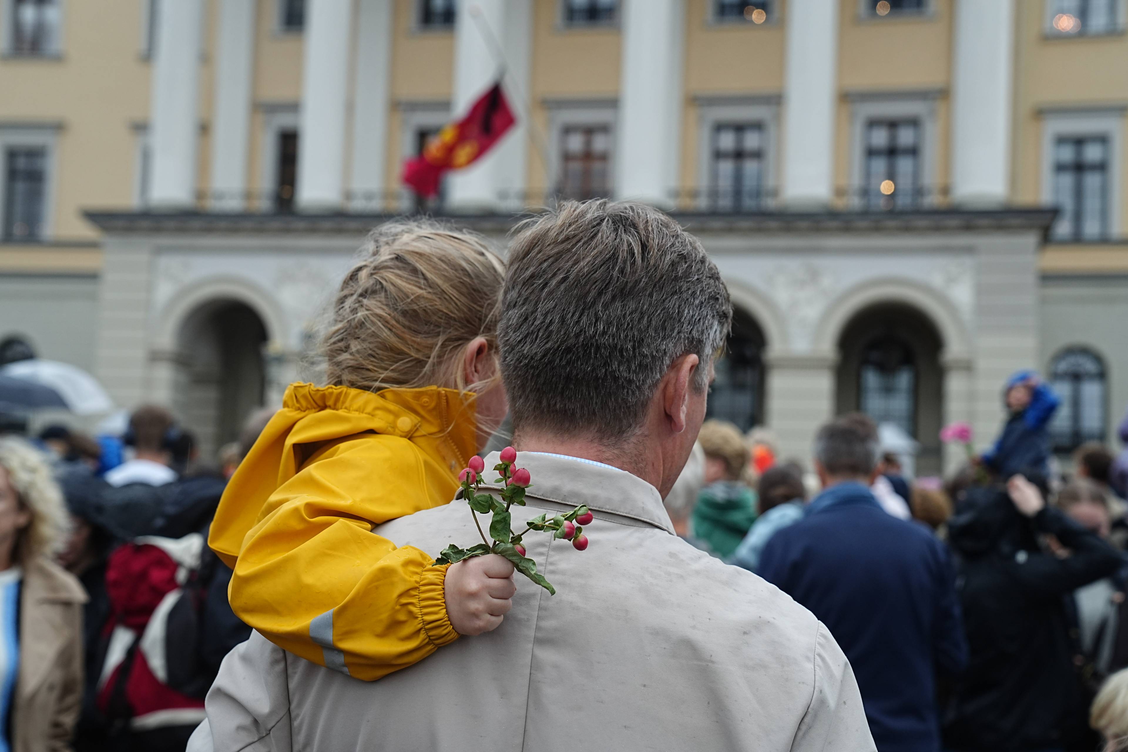En voksen person bærer et barn i gul regnjakke foran Slottet. Barnet holder en kvist med bær. (Bildebeskrivelsen er laget av en KI-tjeneste)
