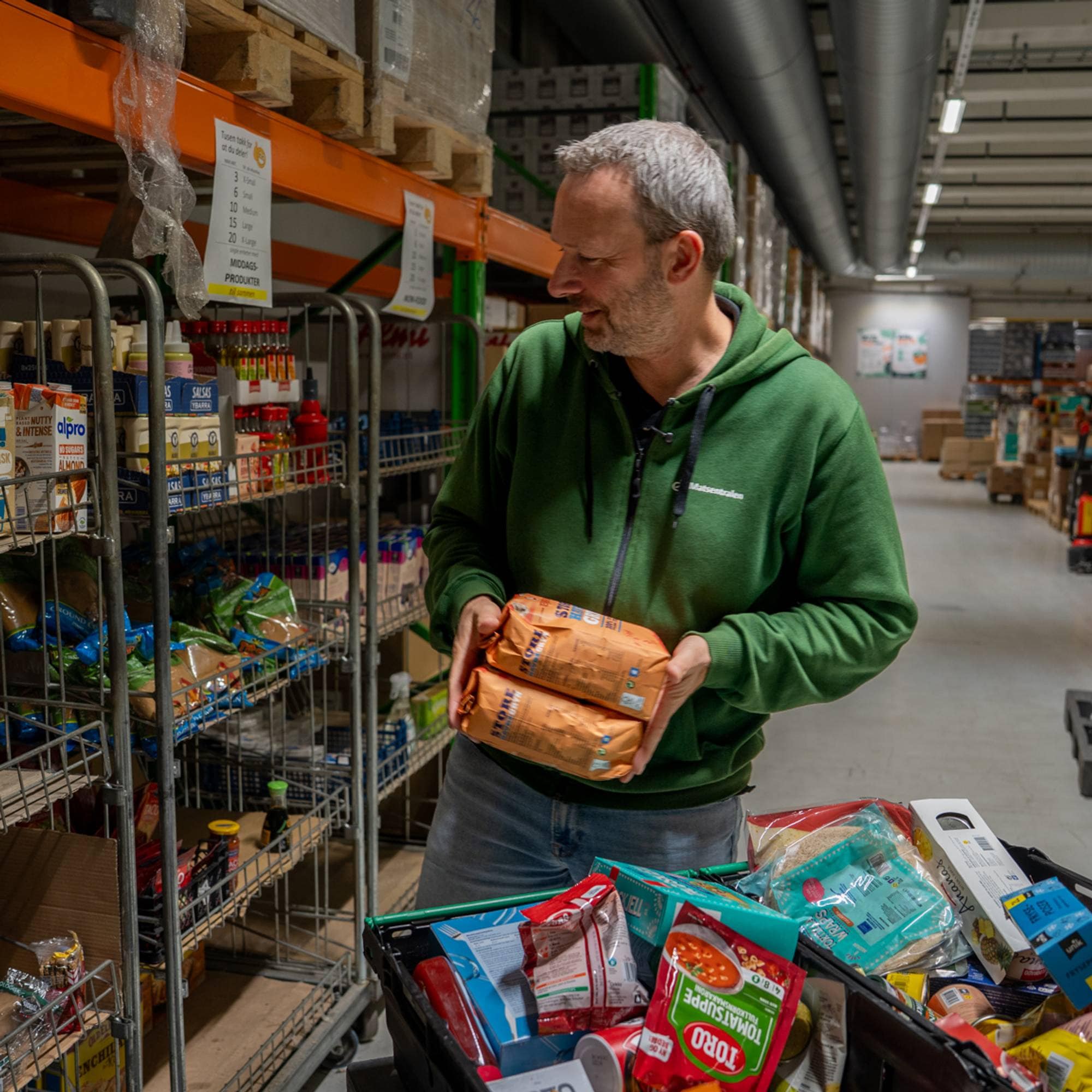 En mann i grønn genser henter matvarer fra hyller på et lager tilhørende Matsentralen.