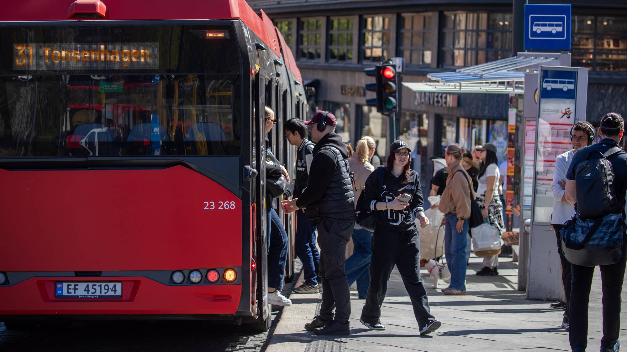 Rød buss med 31 Tonsenhagen står ved en bussholdeplass. Folk går på og av bussen. Flere personer venter på fortauet ved siden av bussholdeplassen. Enkelte har på seg hodetelefoner. (Bildebeskrivelsen er laget av en KI-tjeneste)