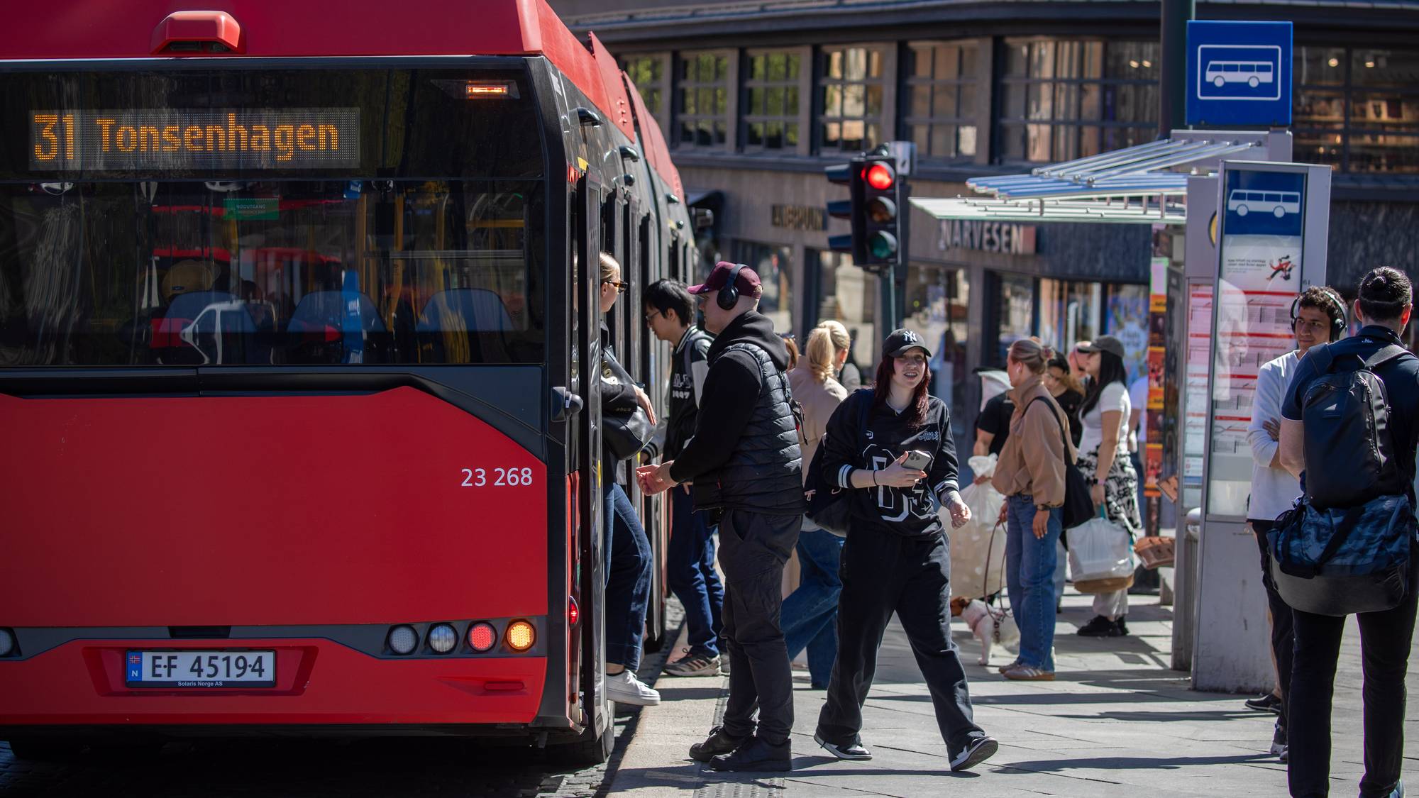 Rød buss med 31 Tonsenhagen står ved en bussholdeplass. Folk går på og av bussen. Flere personer venter på fortauet ved siden av bussholdeplassen. Enkelte har på seg hodetelefoner. (Bildebeskrivelsen er laget av en KI-tjeneste)
