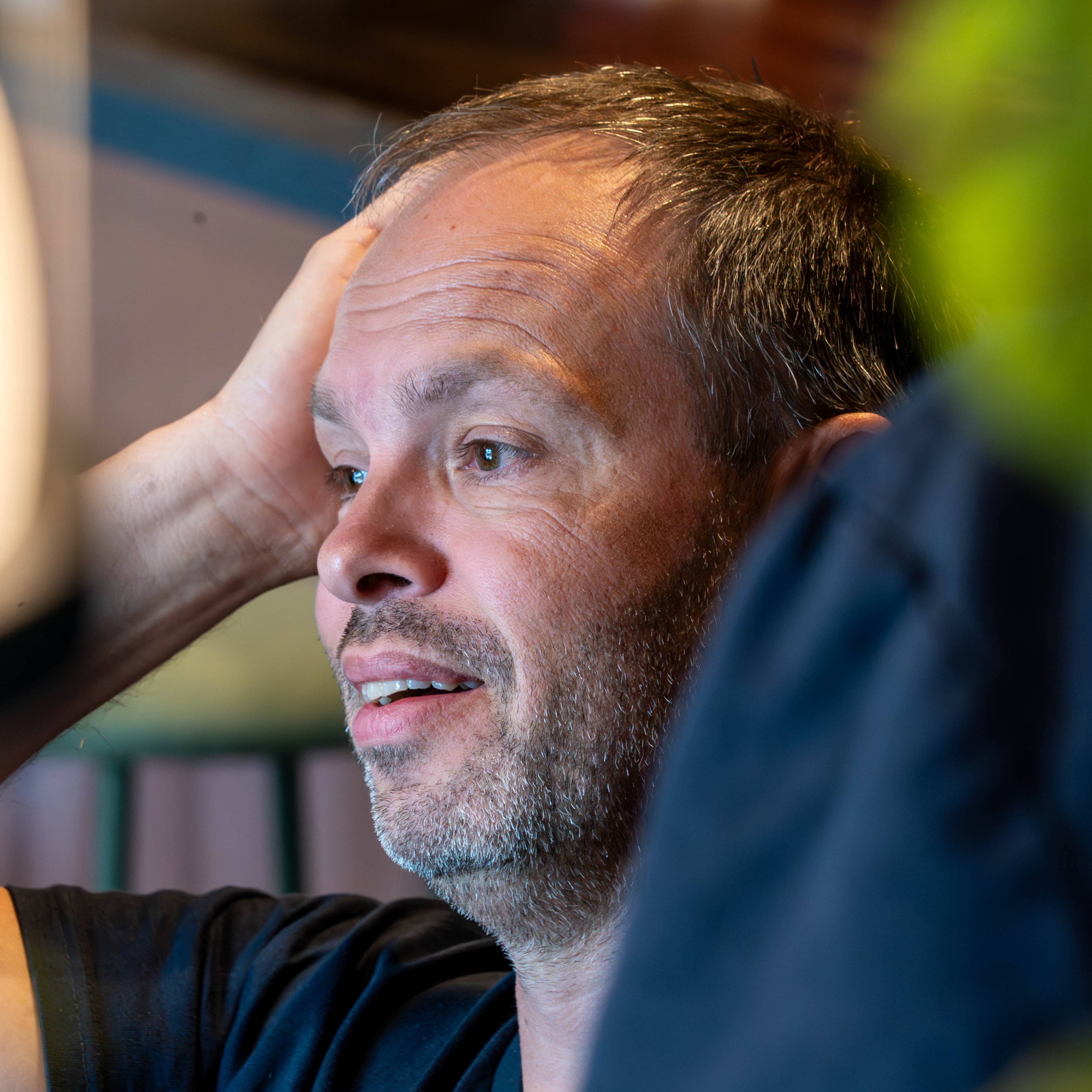 Nærbilde av en mann med kort, grånende hår. Han sitter inne og holder høyre hånd mot hodet. Ansiktet hans er vendt mot venstre, og han ser ut av bildet. En person i mørk skjorte og grønne blader dekker deler av bildet. (Bildebeskrivelsen er laget av en KI-tjeneste)