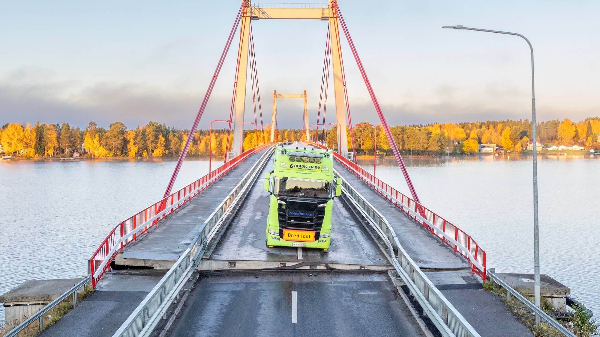 En lastebil kjører over en rød hengebru over en innsjø i høstlandskap. (Bildebeskrivelsen er laget av en KI-tjeneste)