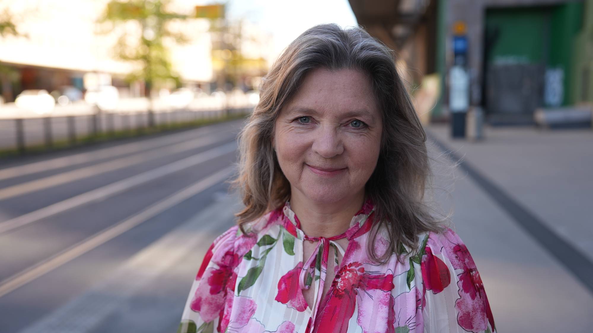 Et nærbilde viser en smilende kvinne som ser mot kamera. Hun har grått og blondt hår og er kledd i en blomstrete bluse. Bak henne ses en gate med trikkeskinner. (Bildebeskrivelsen er laget av en KI-tjeneste)