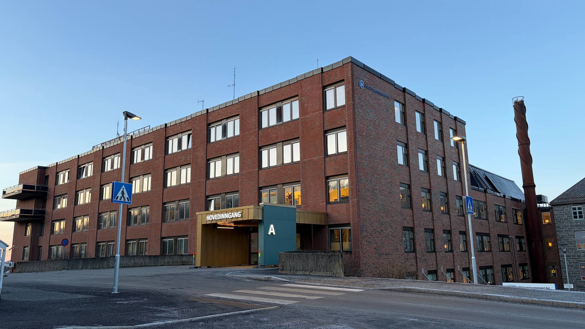 Bygningen i bildet er en modernisert teglstensstruktur med flere vinduer og en hovedinngang merket "HOVEDINNGANG". Den er plassert ved en vei med gangfelt og trafikkskilt. I bakgrunnen ses en høy rød røykrør samt en mindre bygning i mørk stein. Området er ryddig, med klar blå himmel og snø på bakken, noe som gir inntrykk av vinterforhold.