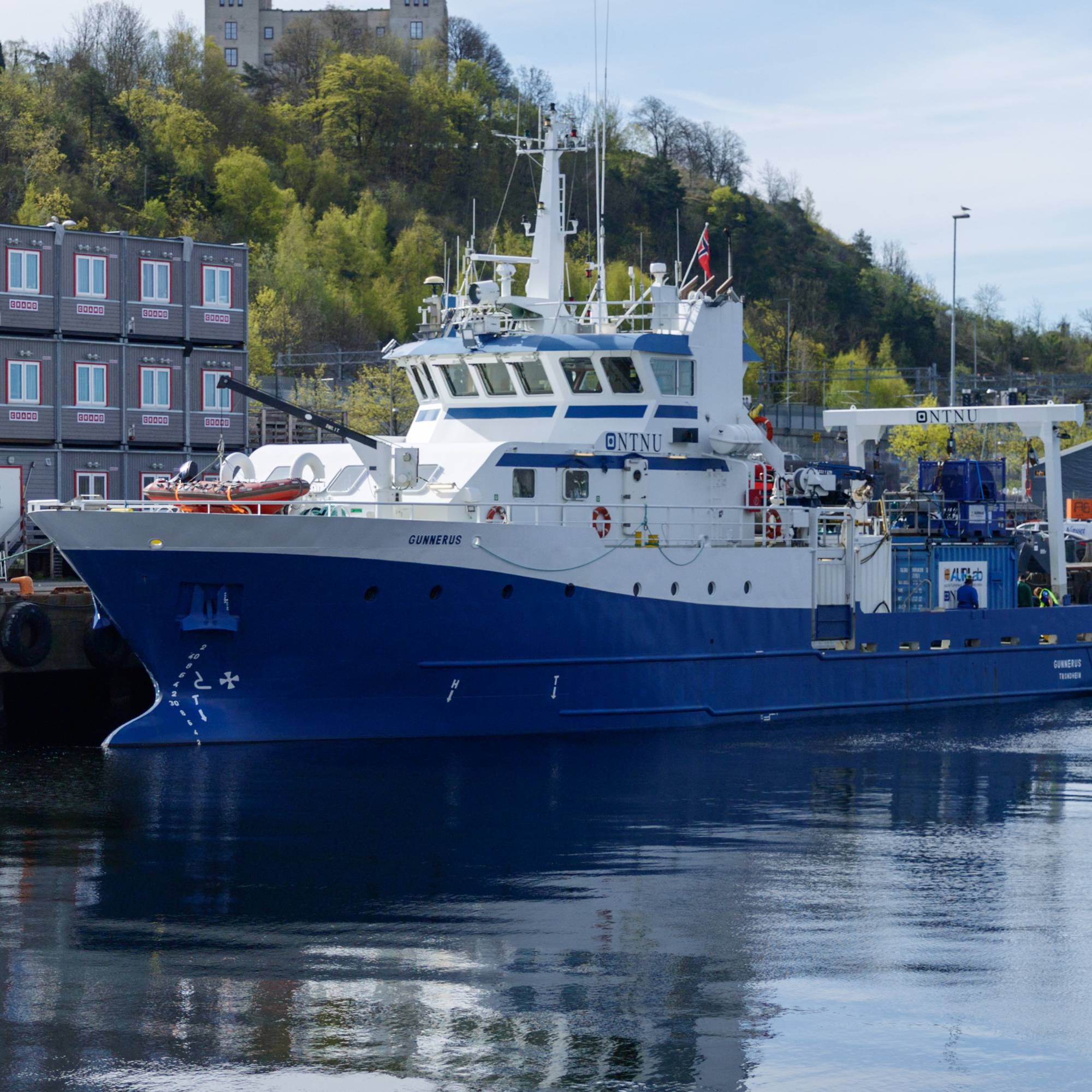 Et blått og hvitt forskningsfartøy ligger ved kai i en havn. Bak fartøyet ligger en lang rekke med containere stablet i høyden. I bakgrunnen er en borg eller festning synlig på en grønnkledd ås. (Bildebeskrivelsen er laget av en KI-tjeneste)
