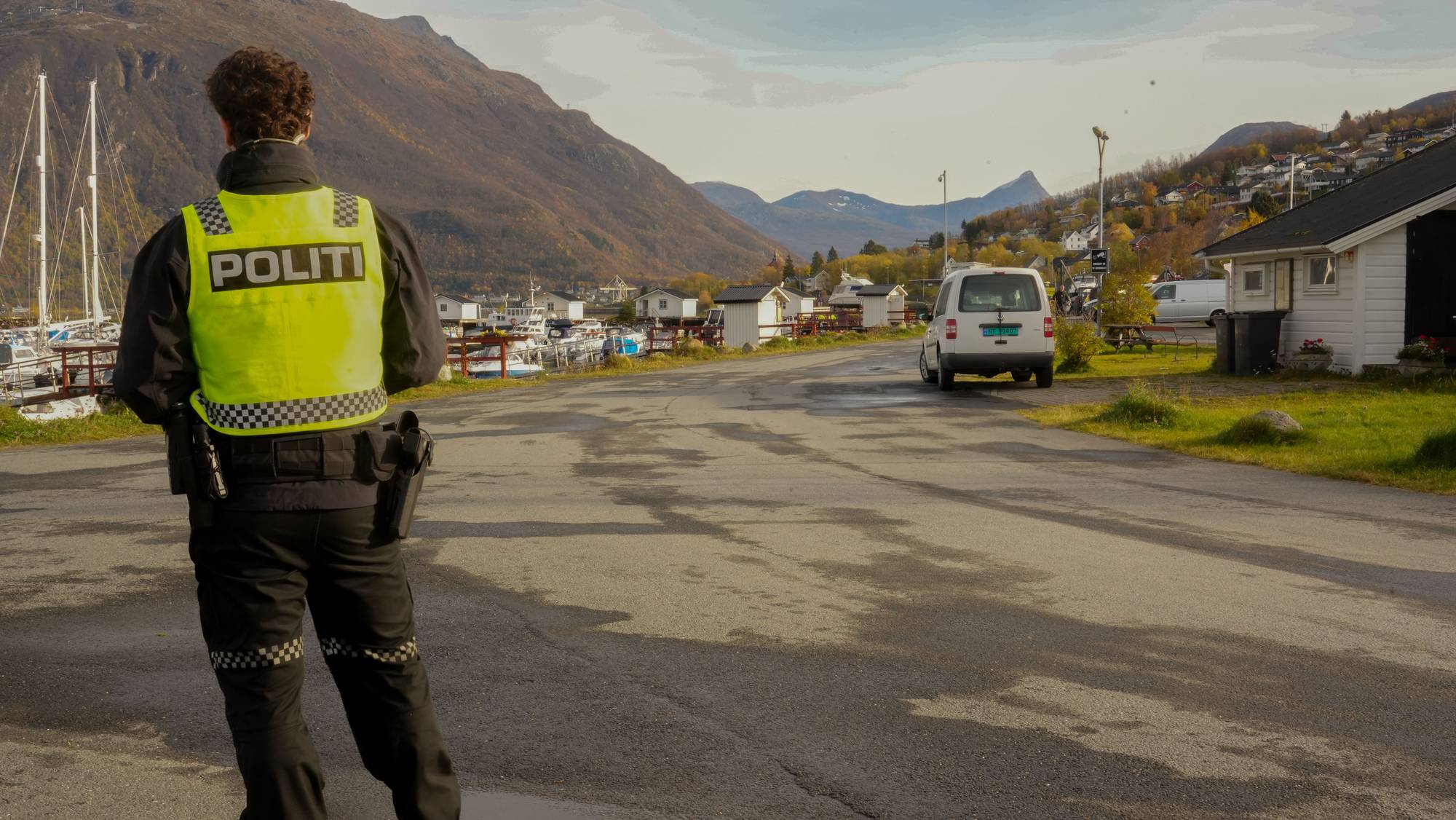 Politibetjent med gul refleksvest ser utover en småbåthavn og et boligområde med fjell i bakgrunnen. (Bildebeskrivelsen er laget av en KI-tjeneste)