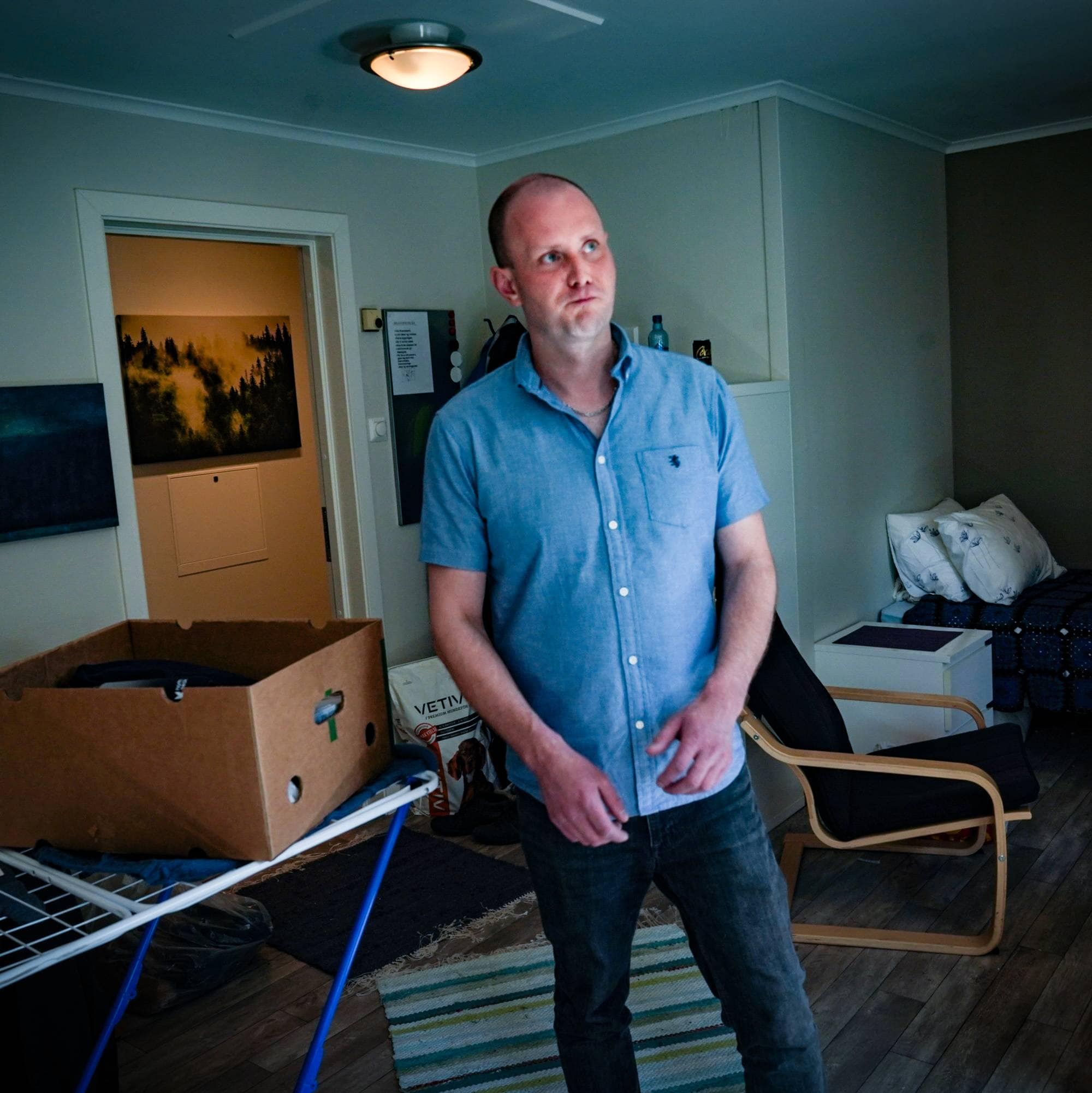 En mann med blå skjorte står midt i et rom. Ved siden av han mpå et tomt tørkestativ står en kasse.