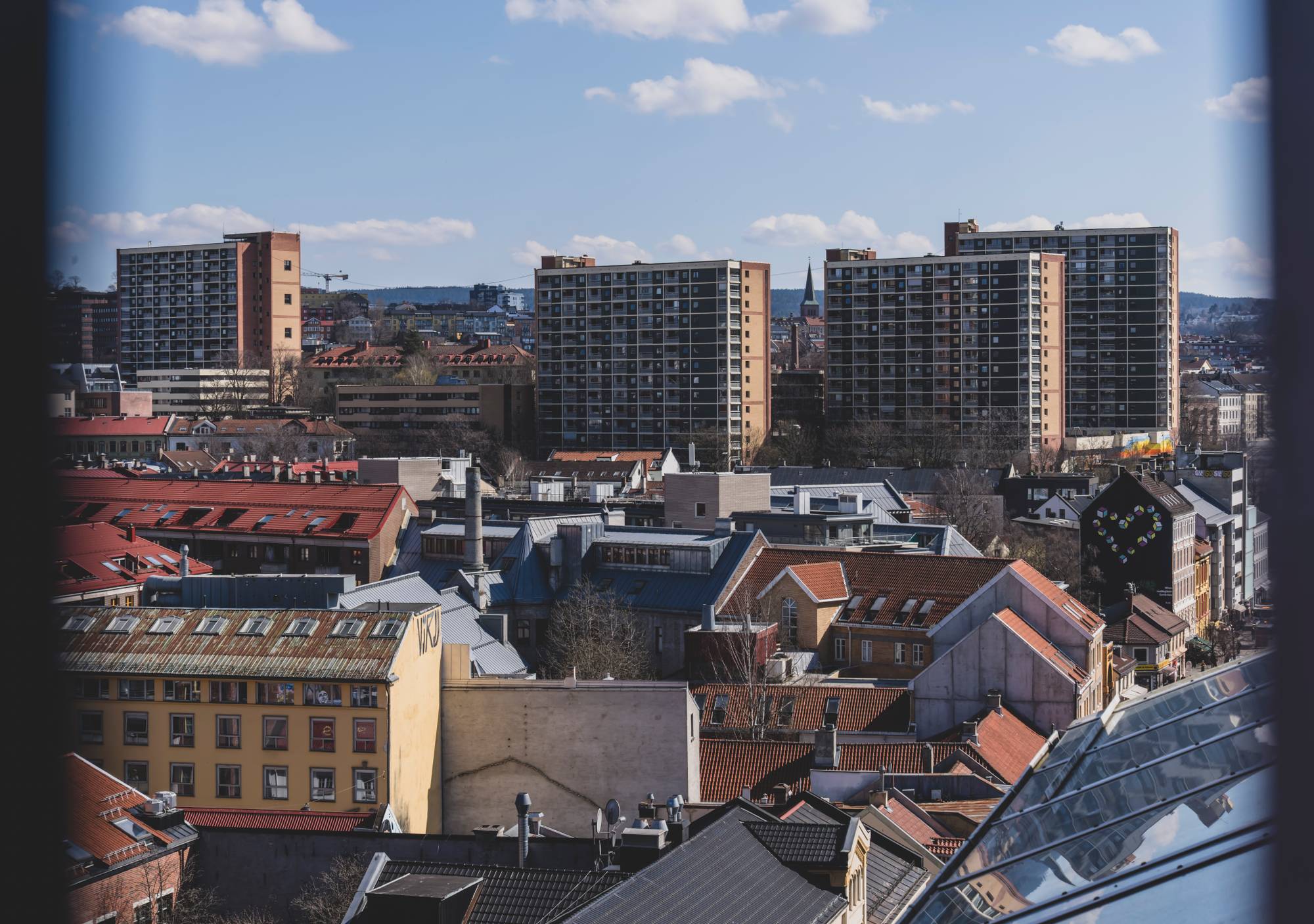 Overblikk over tettbygde boligstrøk med rekkehus og høyblokker. En kirketårn er synlig i bakgrunnen. Klar himmel med noen skyer. Dagen ser ut til å være solrik. (Bildebeskrivelsen er laget av en KI-tjeneste)
