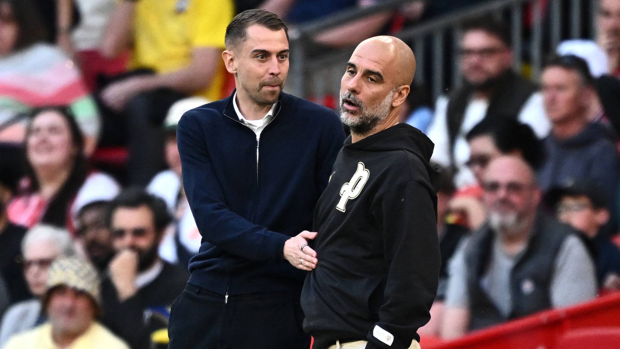 Fotballtrener Pep Guardiola snakker med en kollega på sidelinjen under en kamp foran publikum. (Bildebeskrivelsen er laget av en KI-tjeneste)