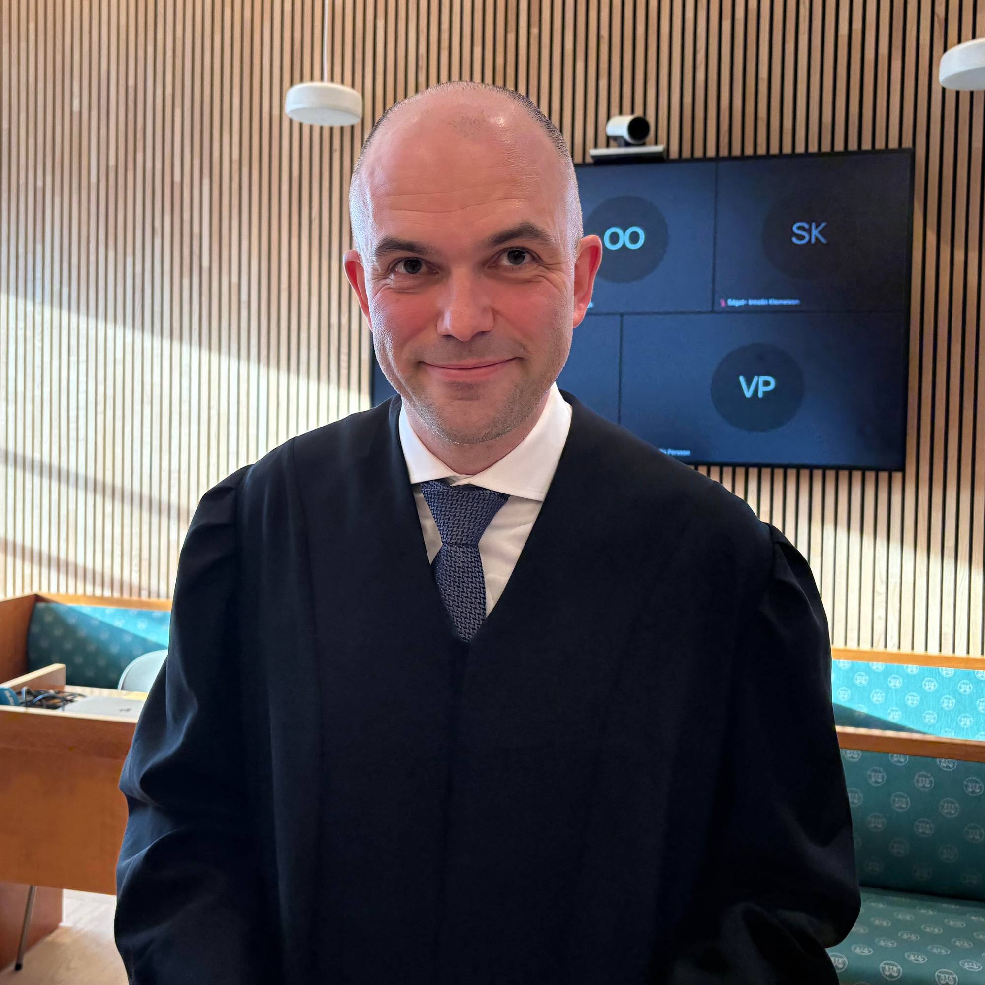 En mann som står i en rettssal. Han har på slips og skjorte. 
