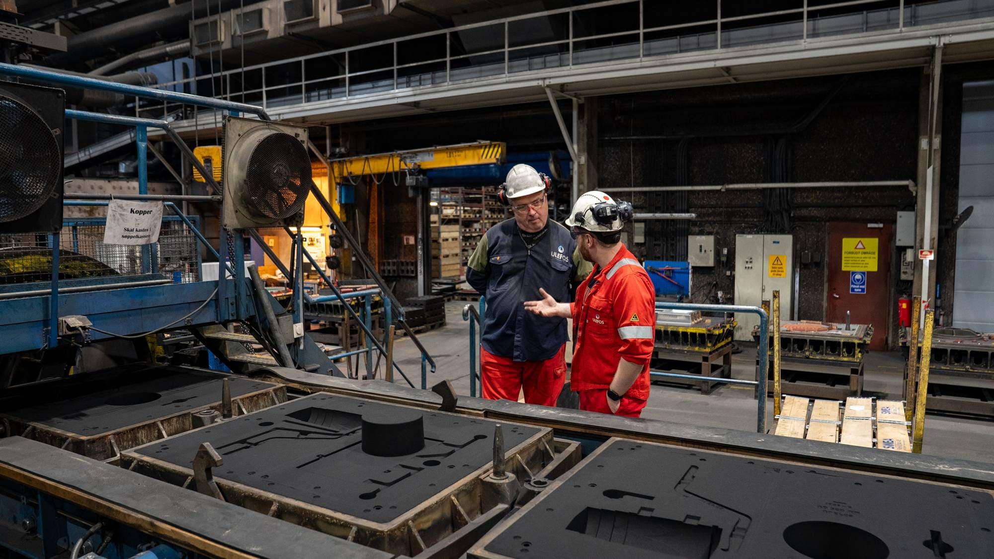 To industriarbeidere i verneutstyr samtaler i en produksjonshall ved støpeformer. (Bildebeskrivelsen er laget av en KI-tjeneste)
