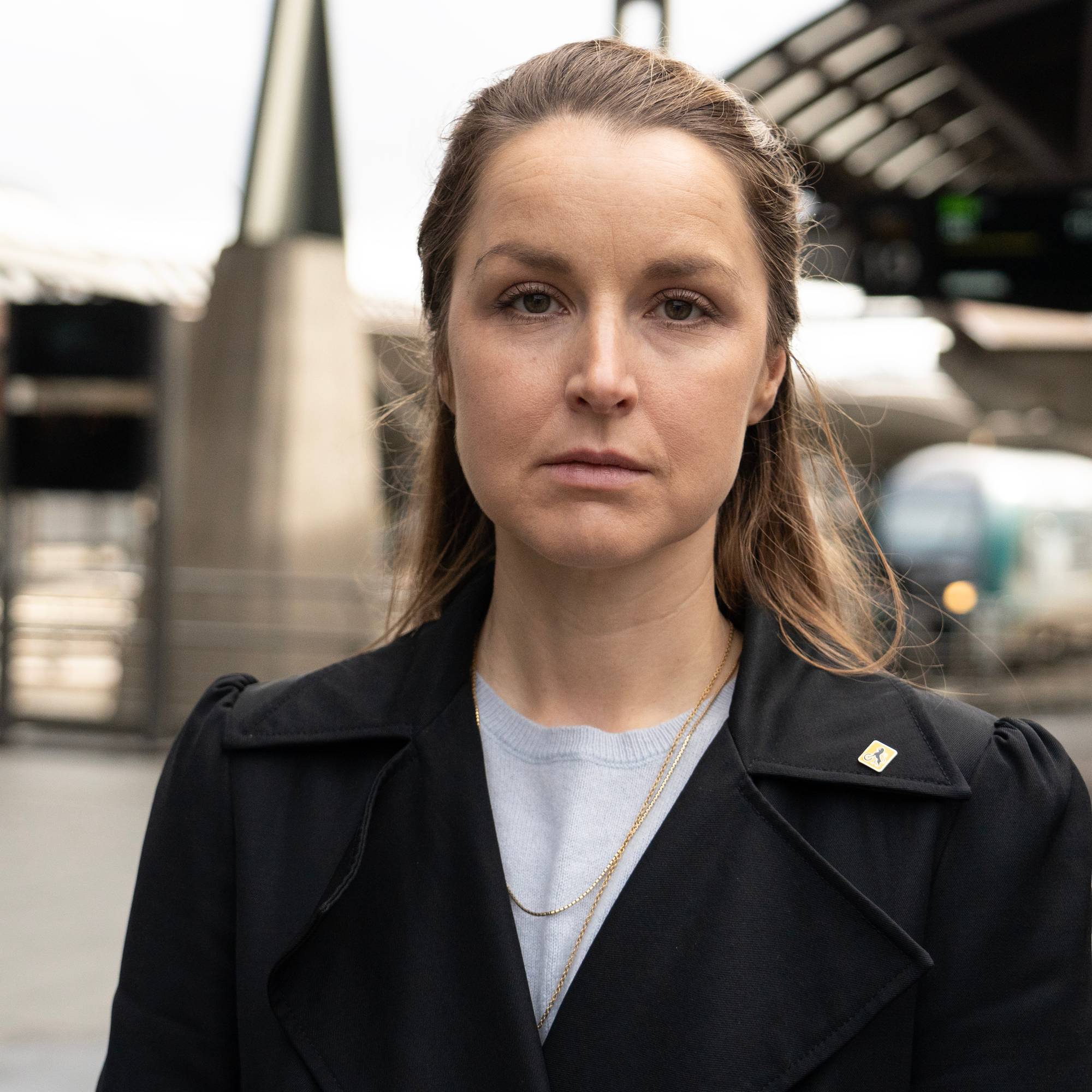 Kvinne med kåpe ser inn i kameraet på en togstasjon. En togvogn er synlig i bakgrunnen. Kvinnen har på seg et smykke og en brosje. (Bildebeskrivelsen er laget av en KI-tjeneste)