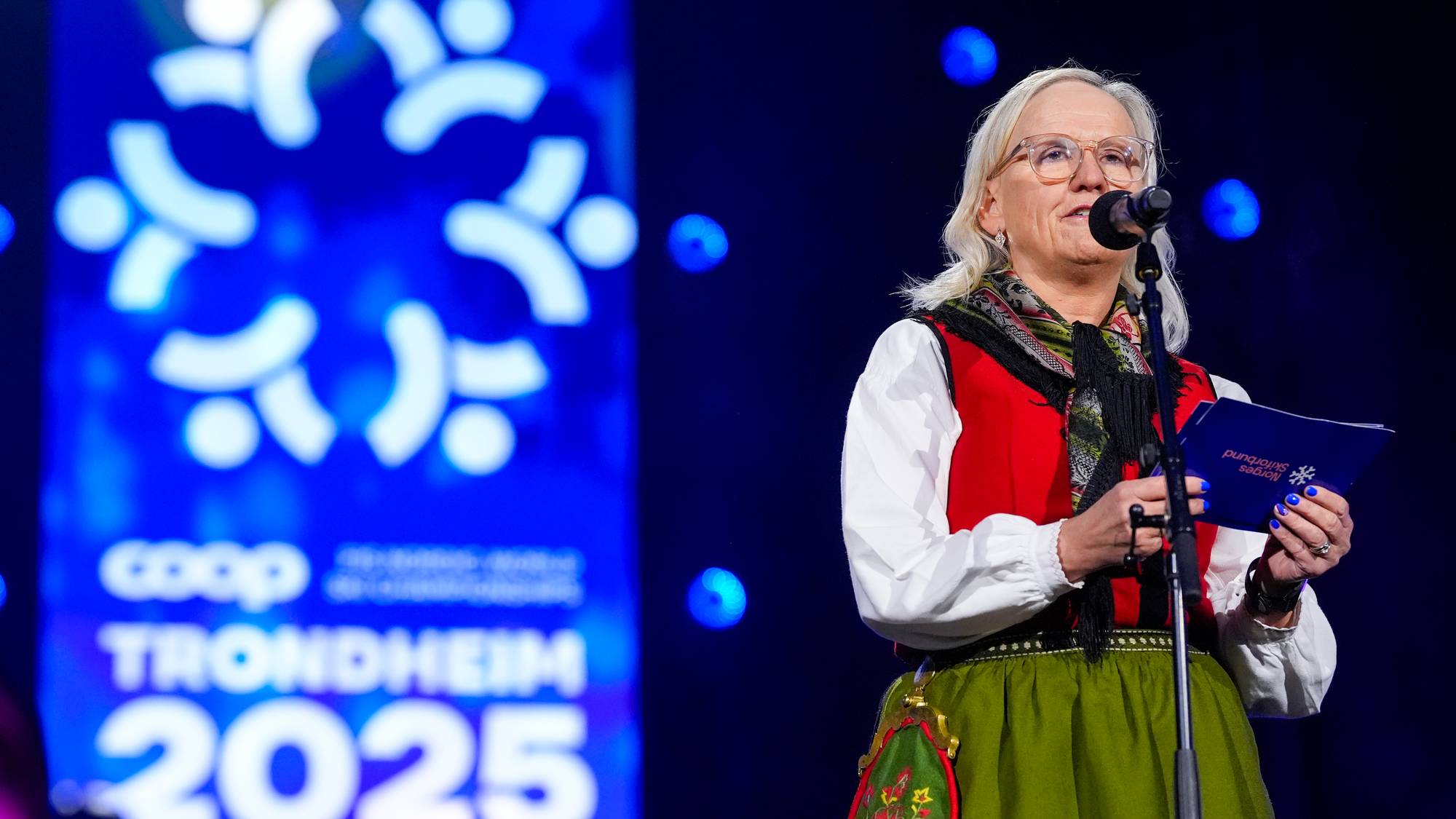 En kvinne i bunad holder et blått hefte og snakker i en mikrofon. Bak henne er en stor, blå bakgrunn med et hvitt logo og teksten «COOP TRONDHEIM 2025». Bakgrunnen har også et blått lysshow. Kvinnen har briller og et skjerf rundt halsen. (Bildebeskrivelsen er laget av en KI-tjeneste)