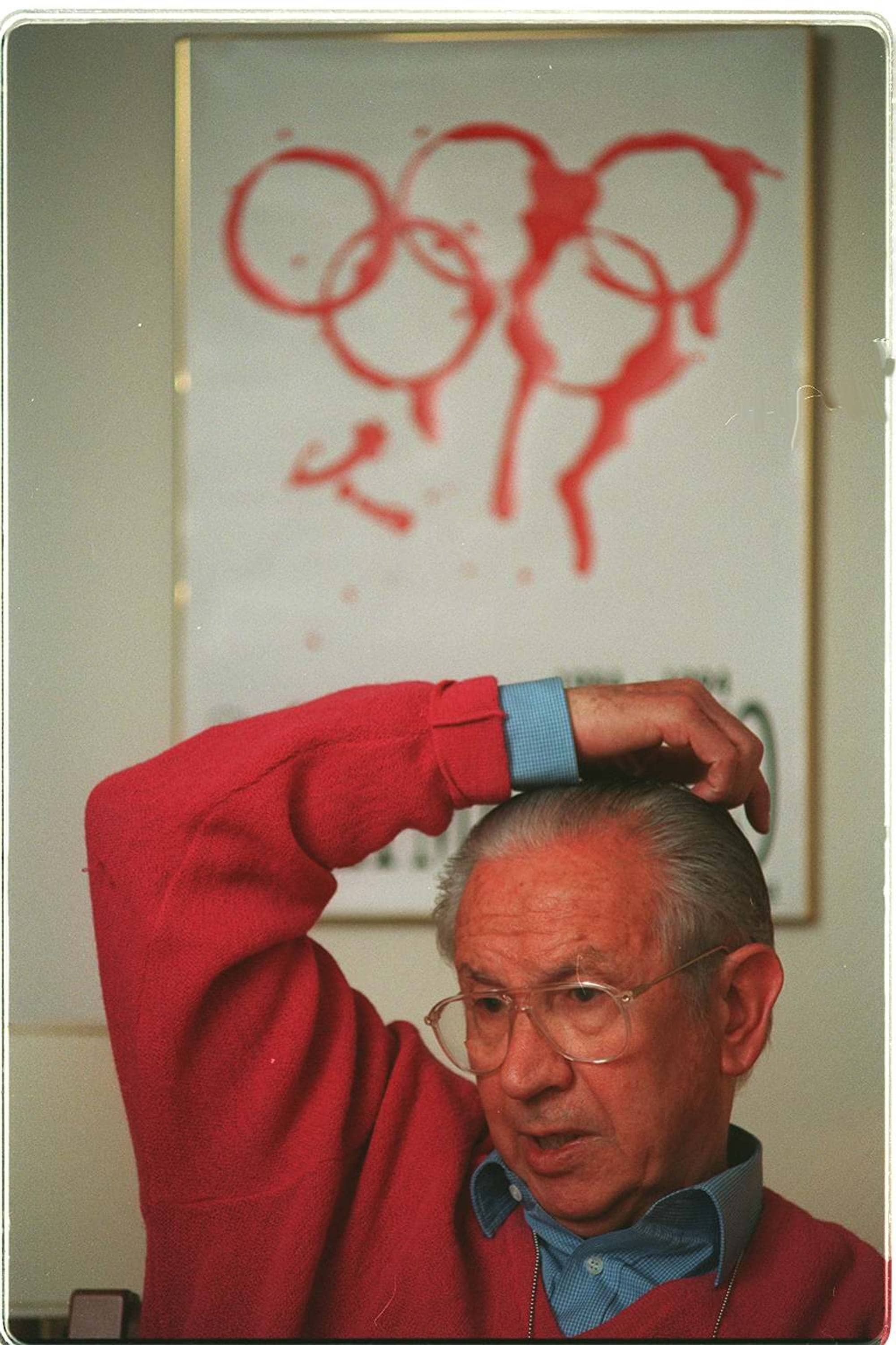 Mann i rød genser foran en plakat med rød olympisk ring-illustrasjon. (Bildebeskrivelsen er laget av en KI-tjeneste)