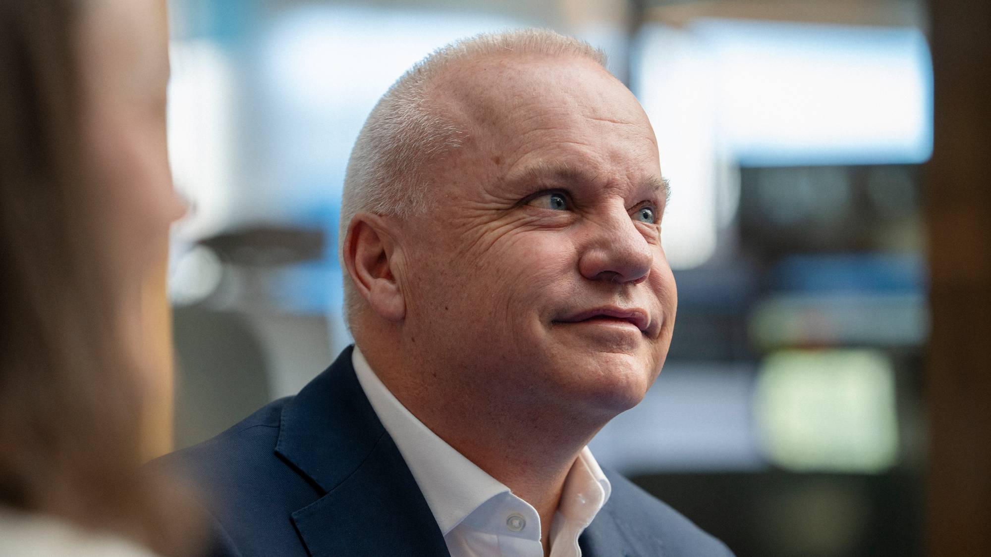 En mann i dress ser til høyre. Bildet er et nærbilde av hans ansikt. Han har kort, grått hår og blå øyne. Mannen er delvis skjult av en person som er ute av fokus til venstre for ham. (Bildebeskrivelsen er laget av en KI-tjeneste)
