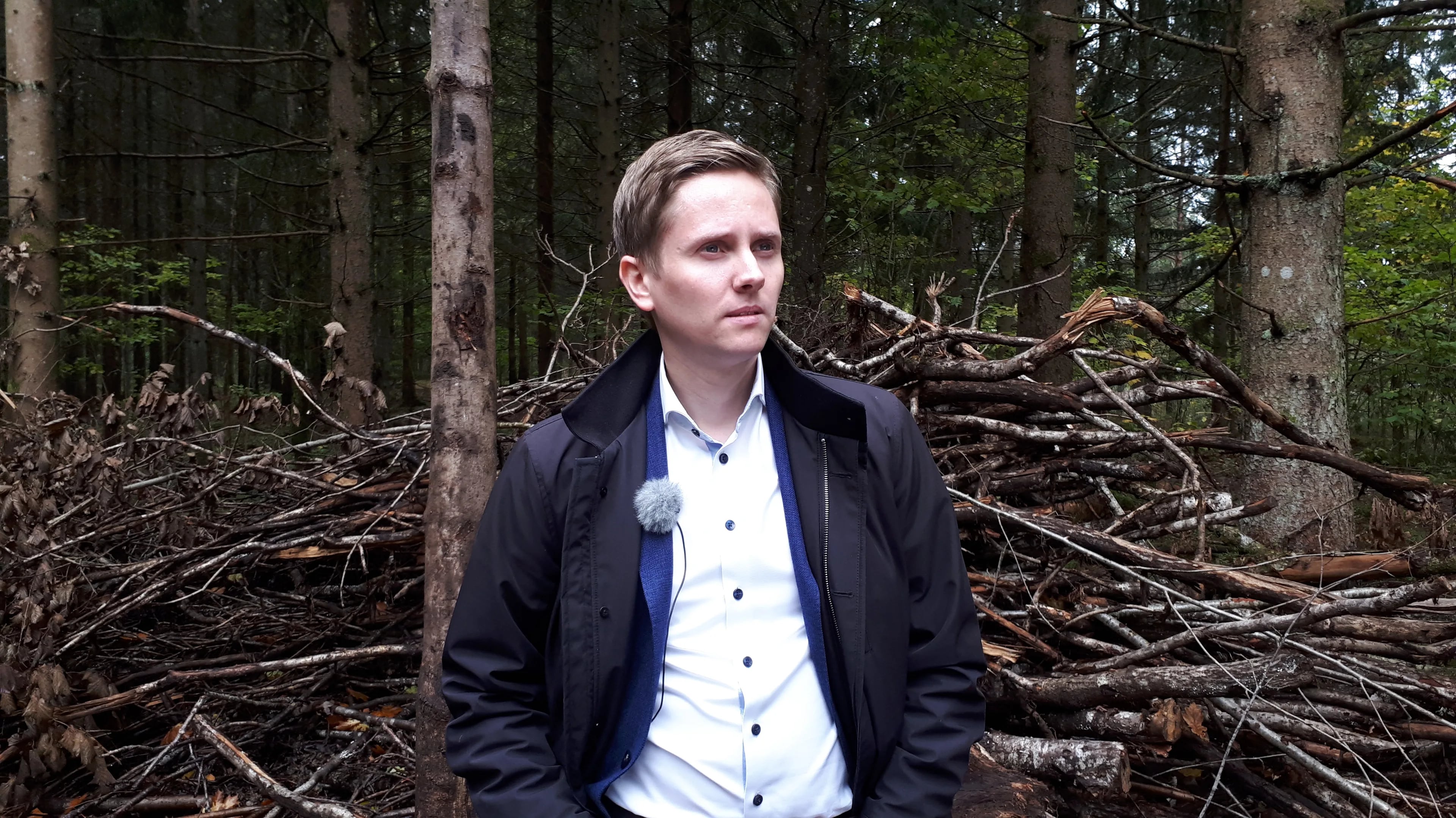 Mann i lys skjorte og mørk jakke med myggmikrofon, stående foran en haug med kvister i en skog. (Bildebeskrivelsen er laget av en KI-tjeneste)