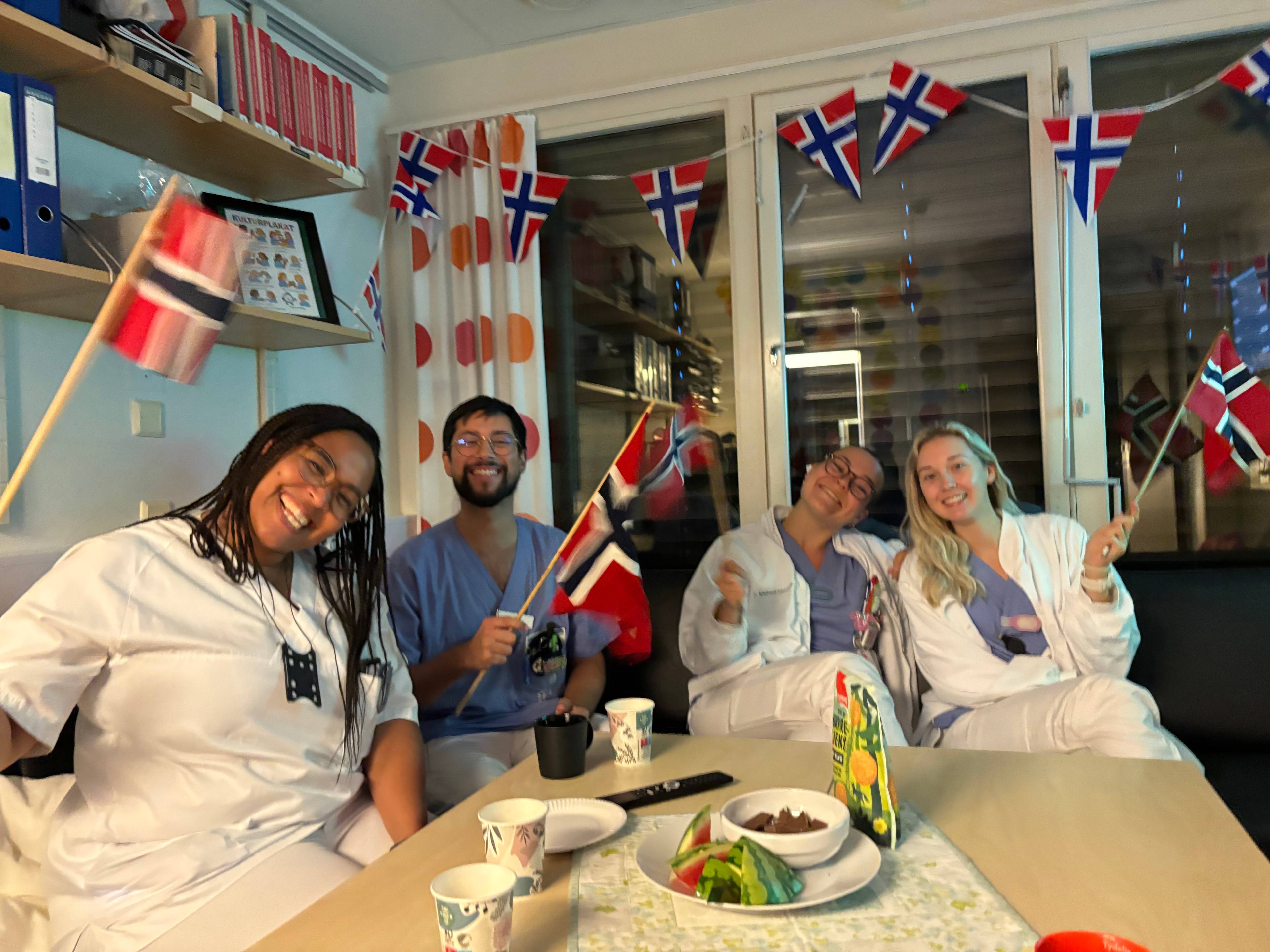 Fire personer, tre kvinner og n mann, sitter ved et bord og holder norske flagg. De har på seg hvite legefrakker og blått arbeidstøy. Veggene er dekorert med norske flagglenker. Personen lengst til venstre smiler bredt mot kameraet mens hun holder et norsk flagg.